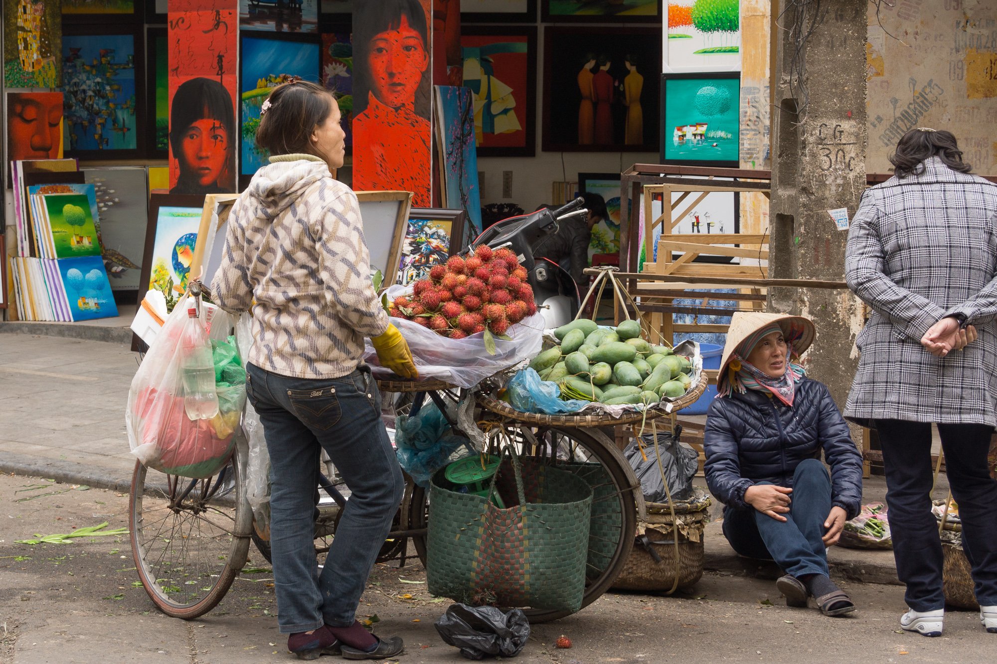 Straßenhändler verkauft rambutan und mango auf einem Fahrrad in einer belebten Straße, im Hintergrund sind farbenfrohe Gemälde in einem Kunstgeschäft.