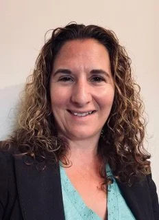 Portrait of a woman with curly hair wearing a teal top and black blazer, smiling in front of a plain background.