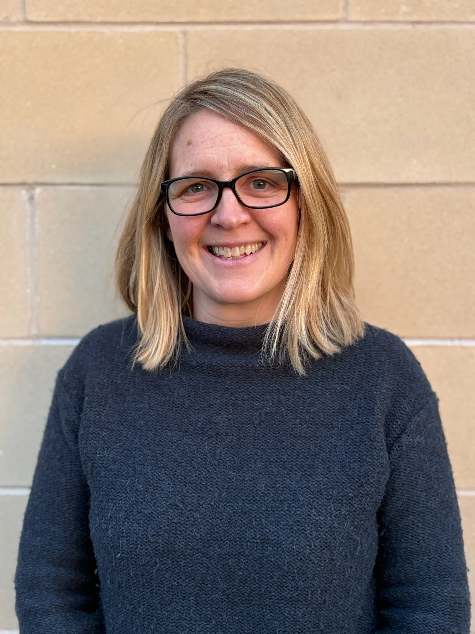 A woman with shoulder-length blond hair wearing a blue denim jacket and a striped shirt, smiling outdoors in front of a stone wall.