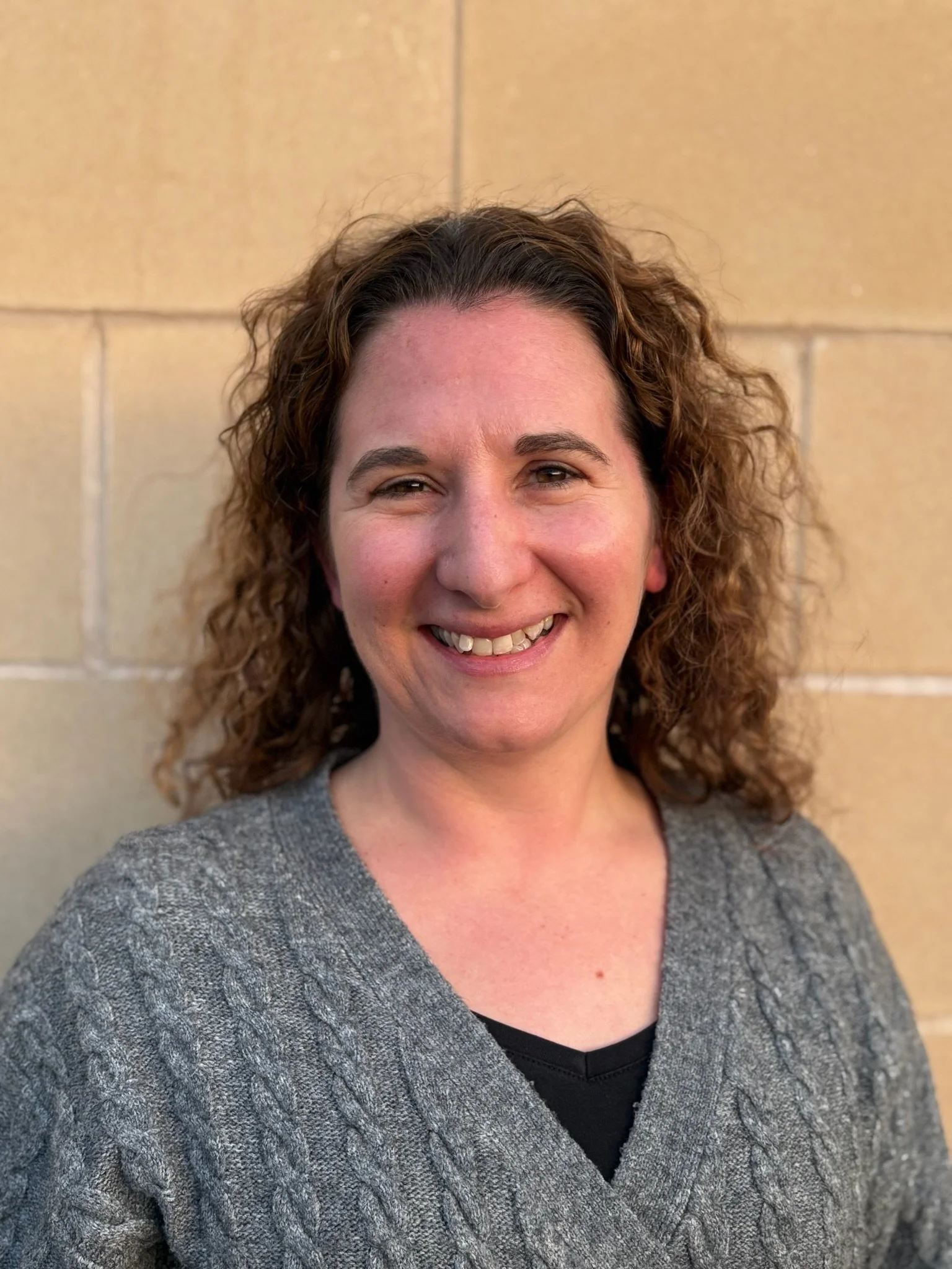 Portrait of a woman with curly hair wearing a teal top and black blazer, smiling in front of a plain background.