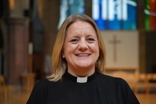 A woman dressed as a priest inside a church, smiling at the camera.