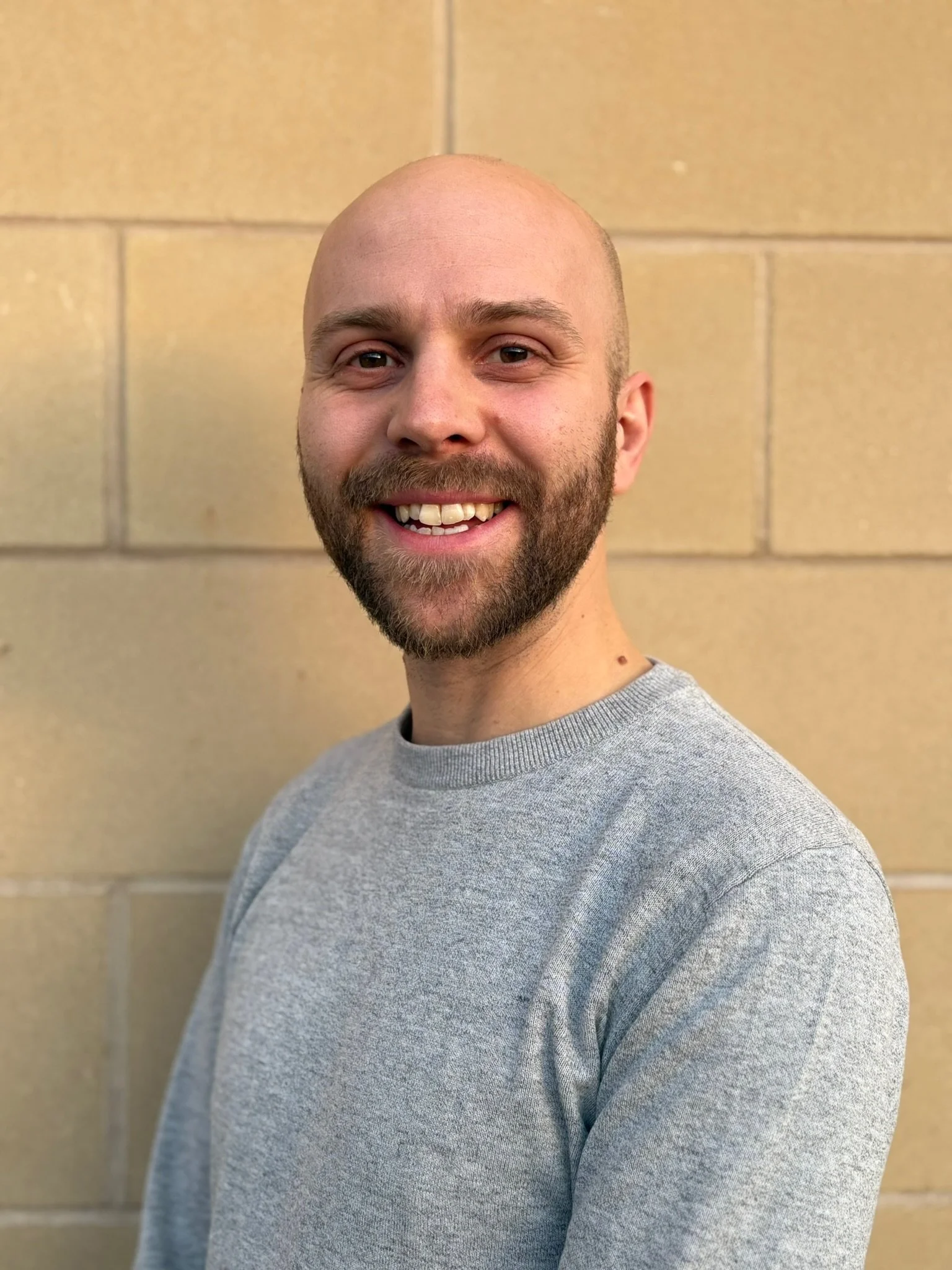 Young man wearing glasses and a brown jacket, outdoors in front of a stone wall.