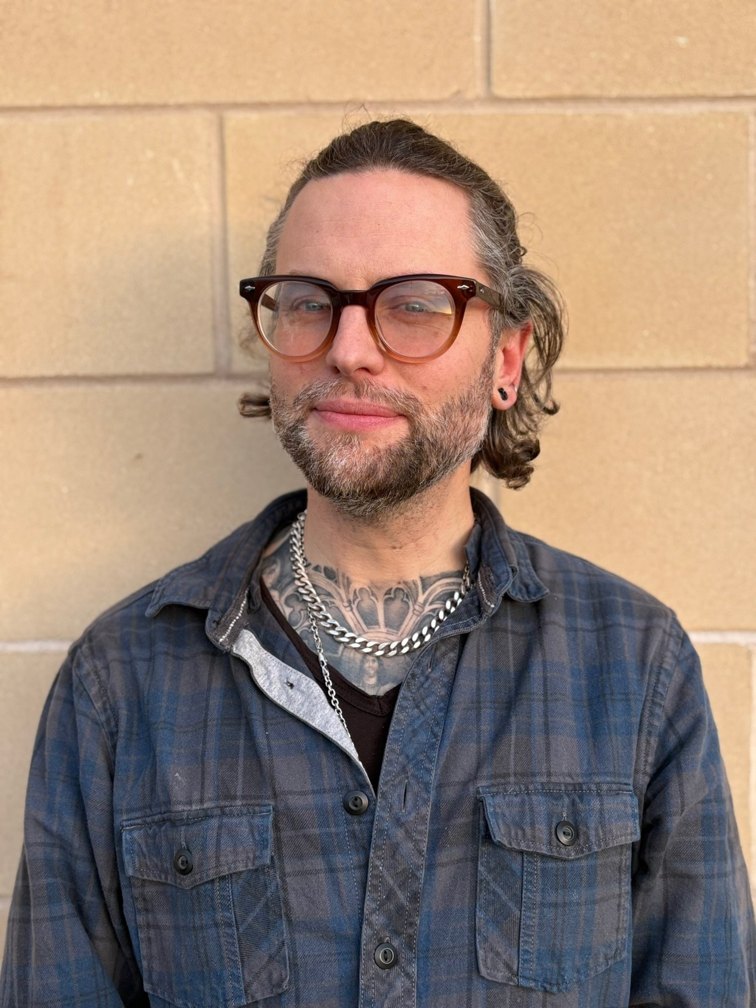 A woman with shoulder-length blond hair wearing a blue denim jacket and a striped shirt, smiling outdoors in front of a stone wall.