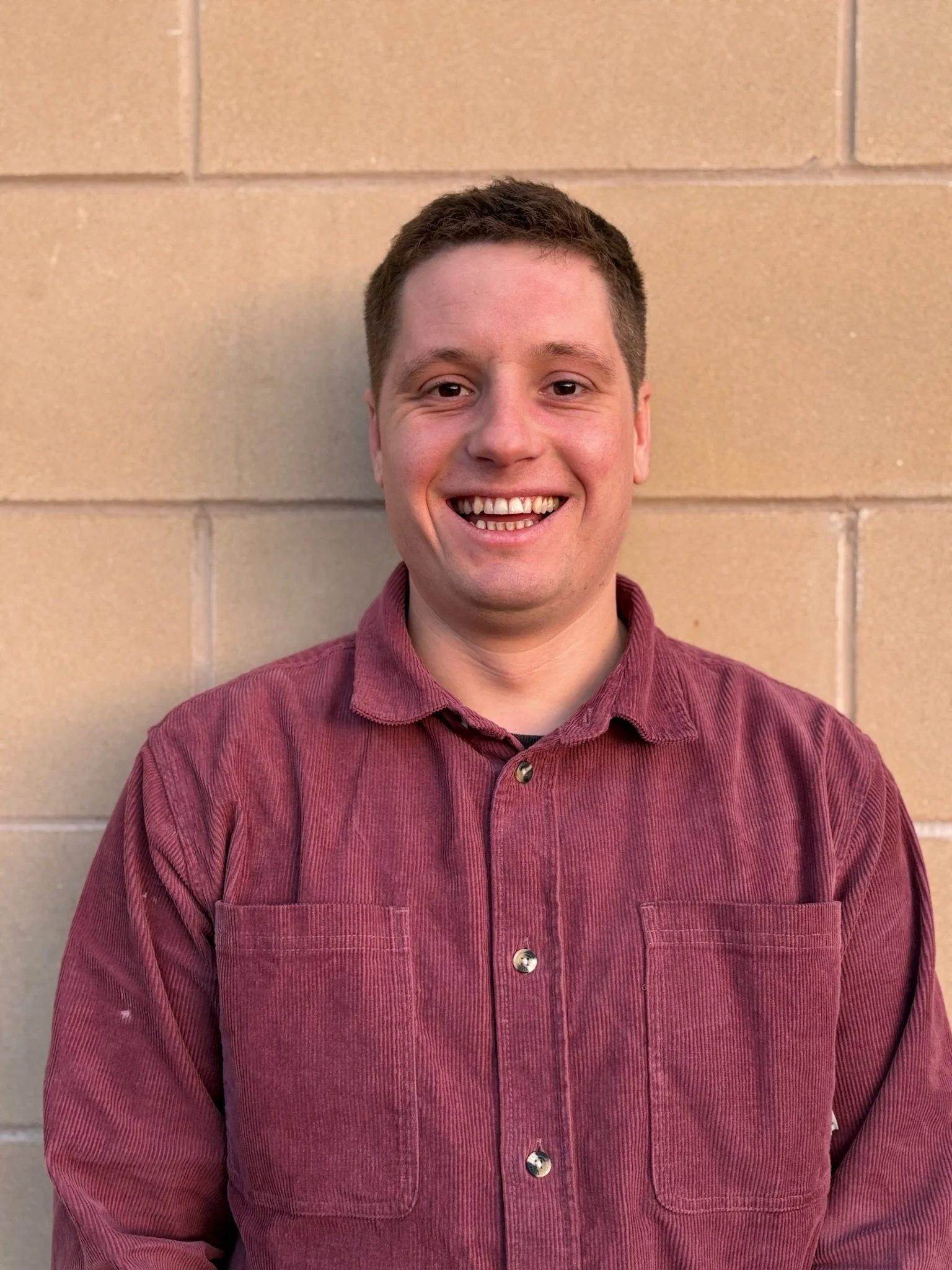 A woman with shoulder-length blond hair wearing a blue denim jacket and a striped shirt, smiling outdoors in front of a stone wall.