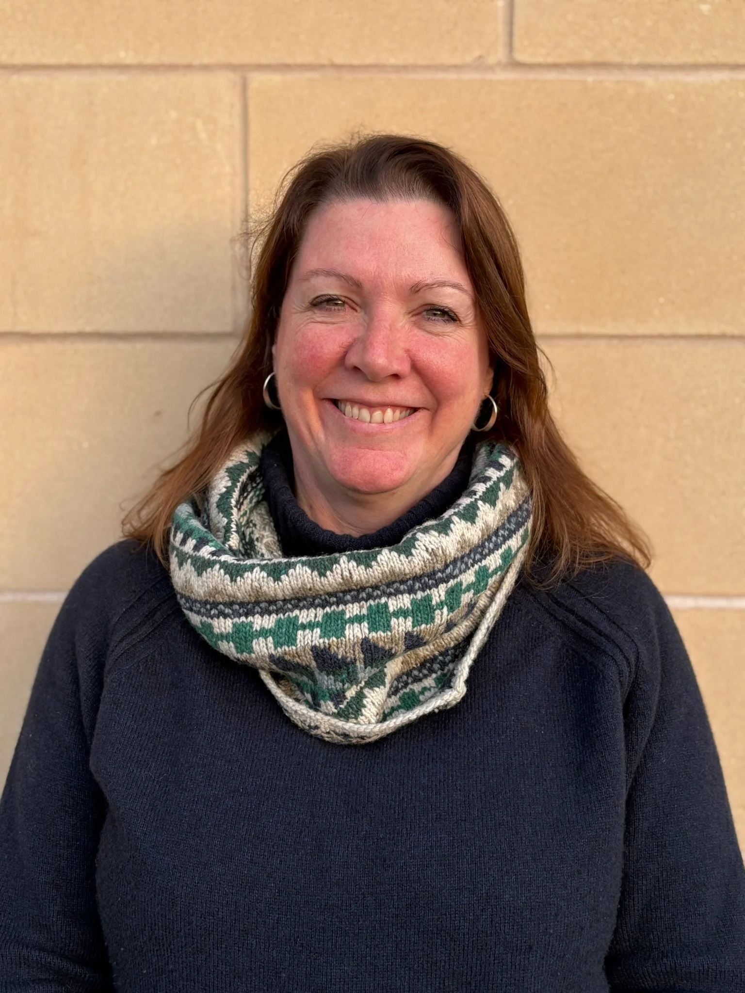Young man wearing glasses and a brown jacket, outdoors in front of a stone wall.