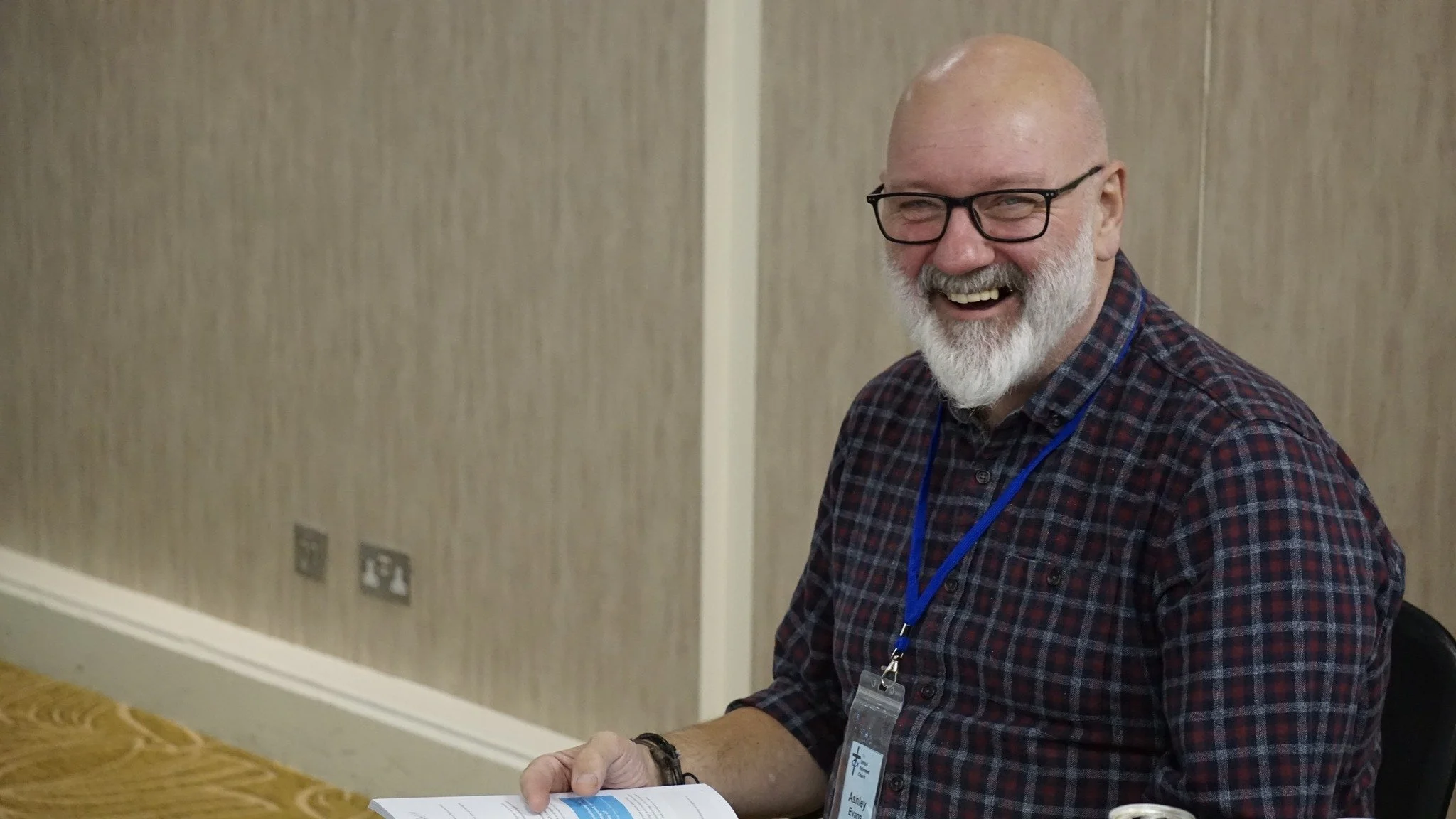 Smiling man with glasses and a beard wearing a plaid shirt and a name badge sitting at a table, holding a booklet, in a conference room.