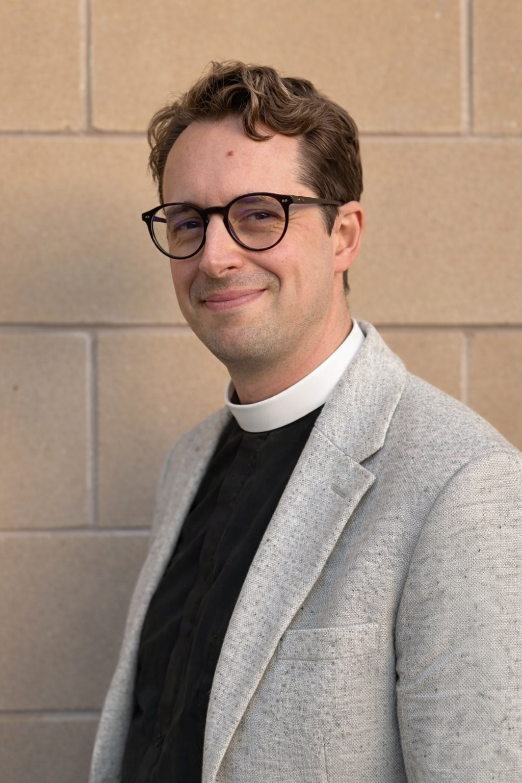 Young man wearing glasses and a brown jacket, outdoors in front of a stone wall.
