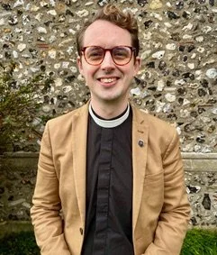 Young man wearing glasses and a brown jacket, outdoors in front of a stone wall.