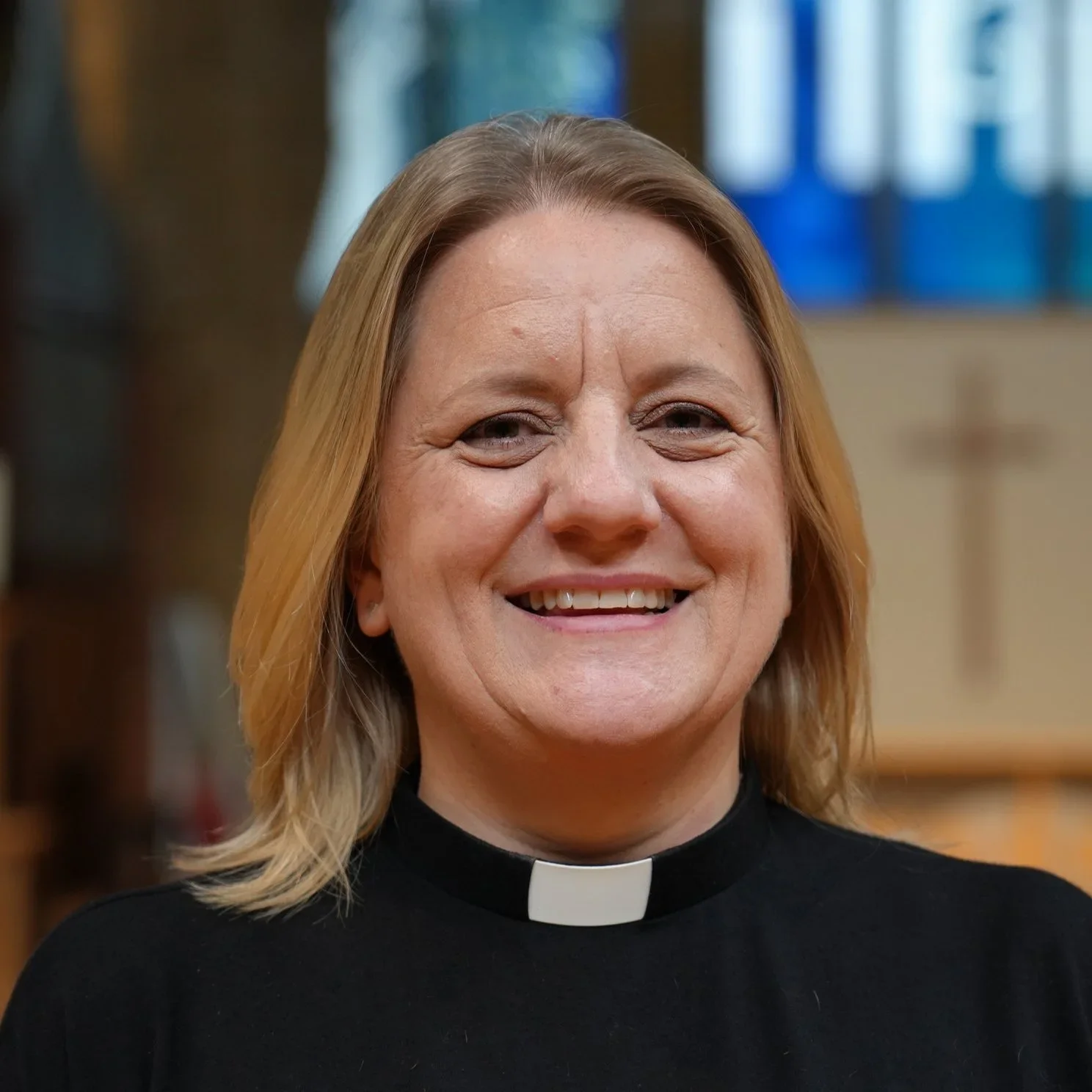 Smiling woman with shoulder-length blonde hair, wearing a black clerical shirt with a white collar, standing inside a church.
