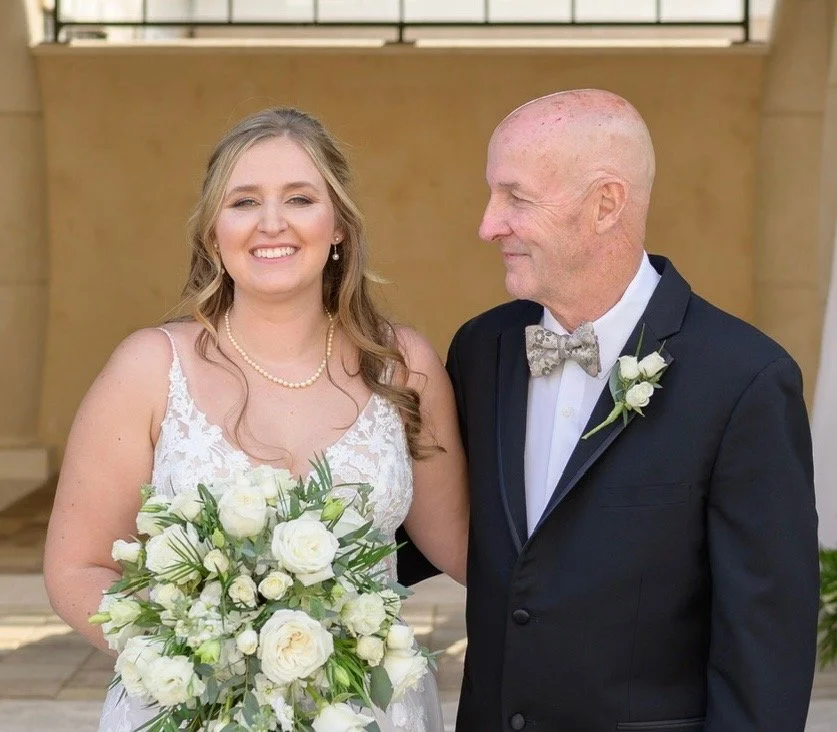 A bride in a white lace wedding dress holding a bouquet of white flowers, standing next to an older man in a black suit with a bow tie and boutonniere, smiling at each other inside a building with beige walls.