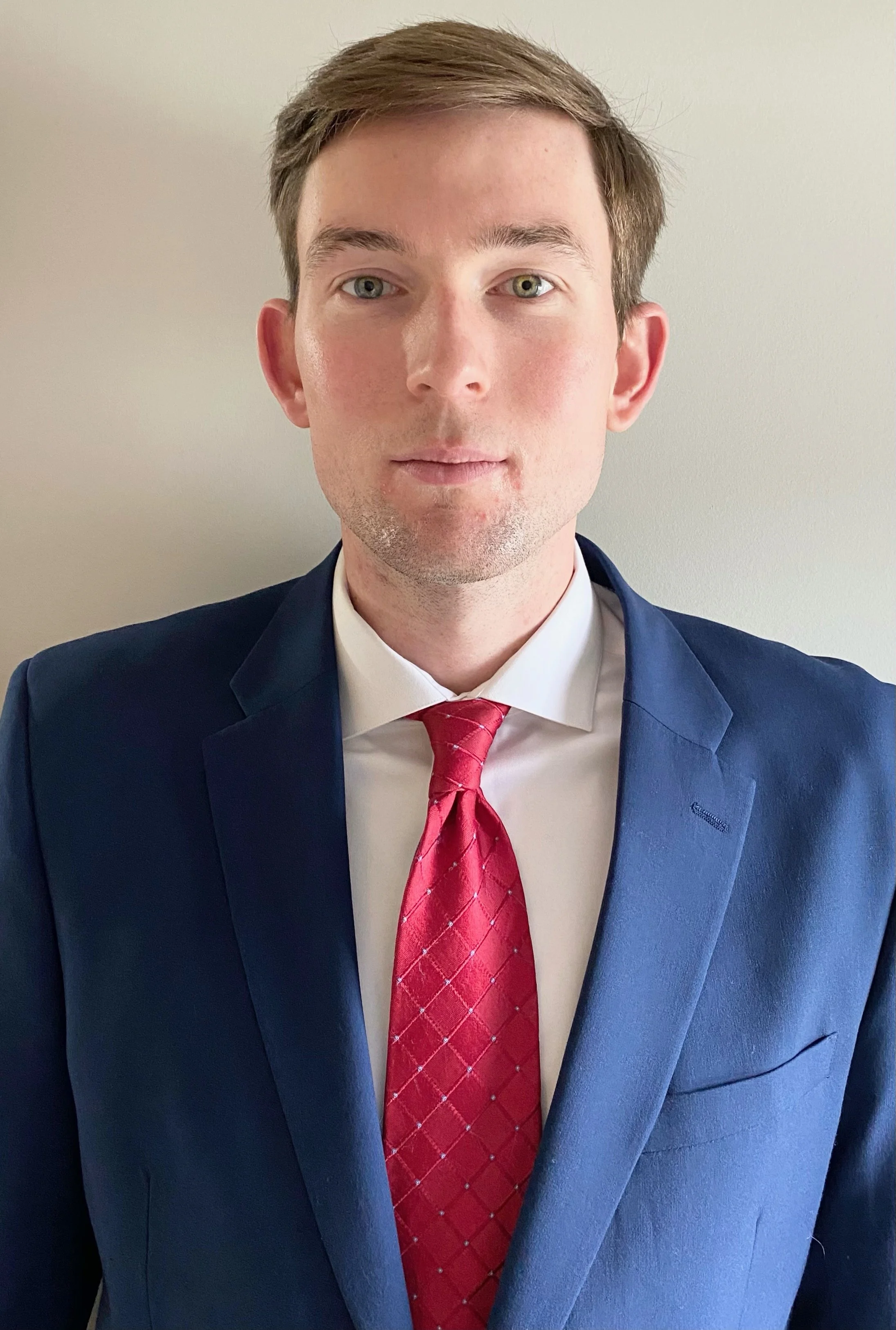 A young man wearing a navy blue suit, white dress shirt, and red tie, posing for a photo against a plain light-colored wall.