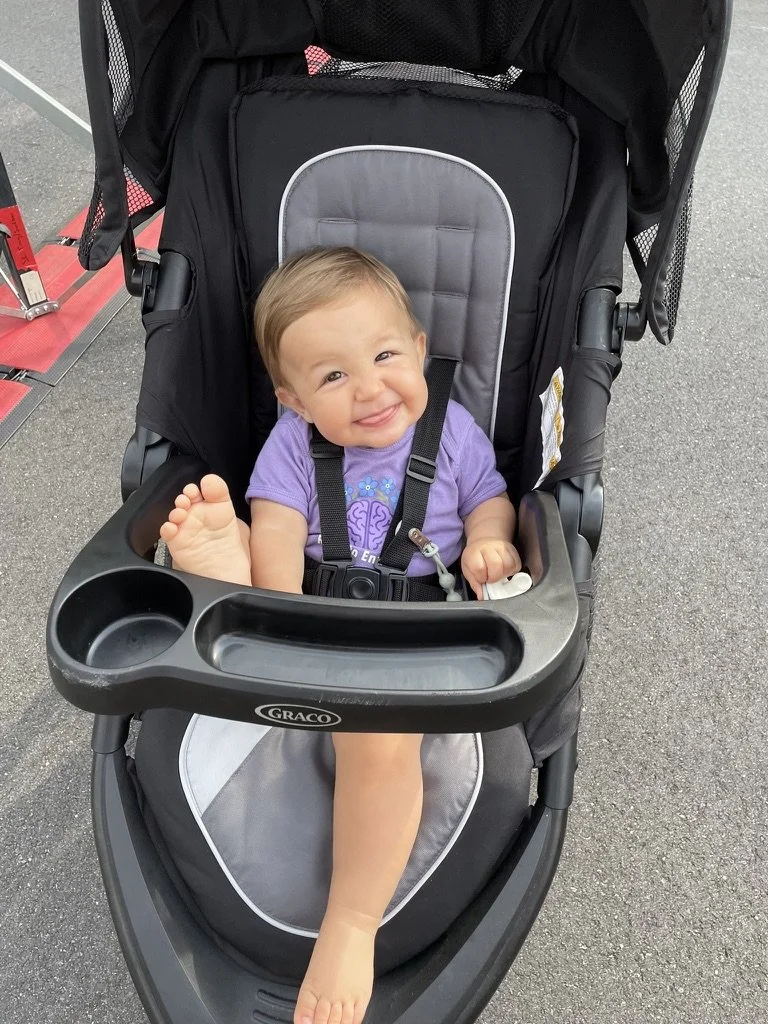 A smiling young child with light brown hair sitting in a black and gray stroller outdoors on a paved surface, wearing a purple shirt, foot resting on the stroller's tray.