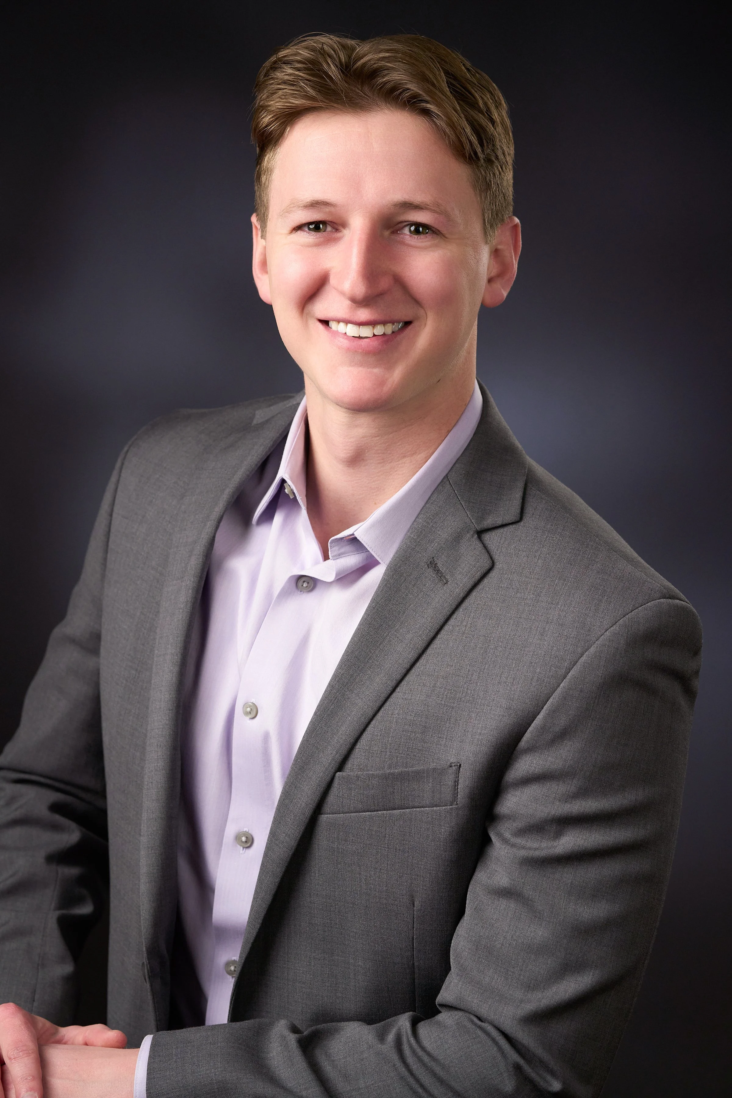 Professional portrait of a young man with light brown hair, wearing a gray business suit and a light purple shirt, smiling against a dark background.