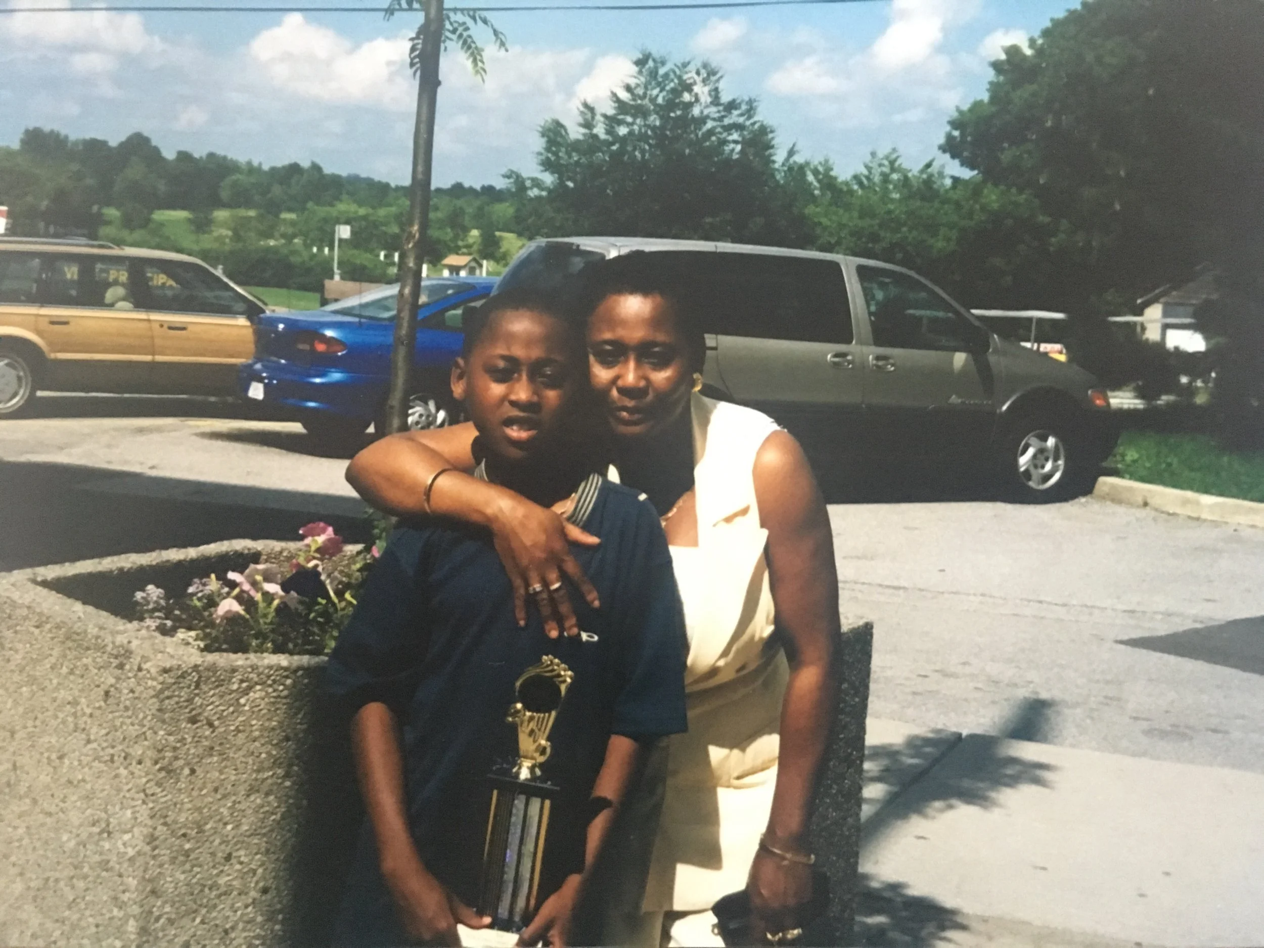 A woman and a boy standing outdoors in a parking lot, with cars and trees in the background. The boy is holding a trophy.