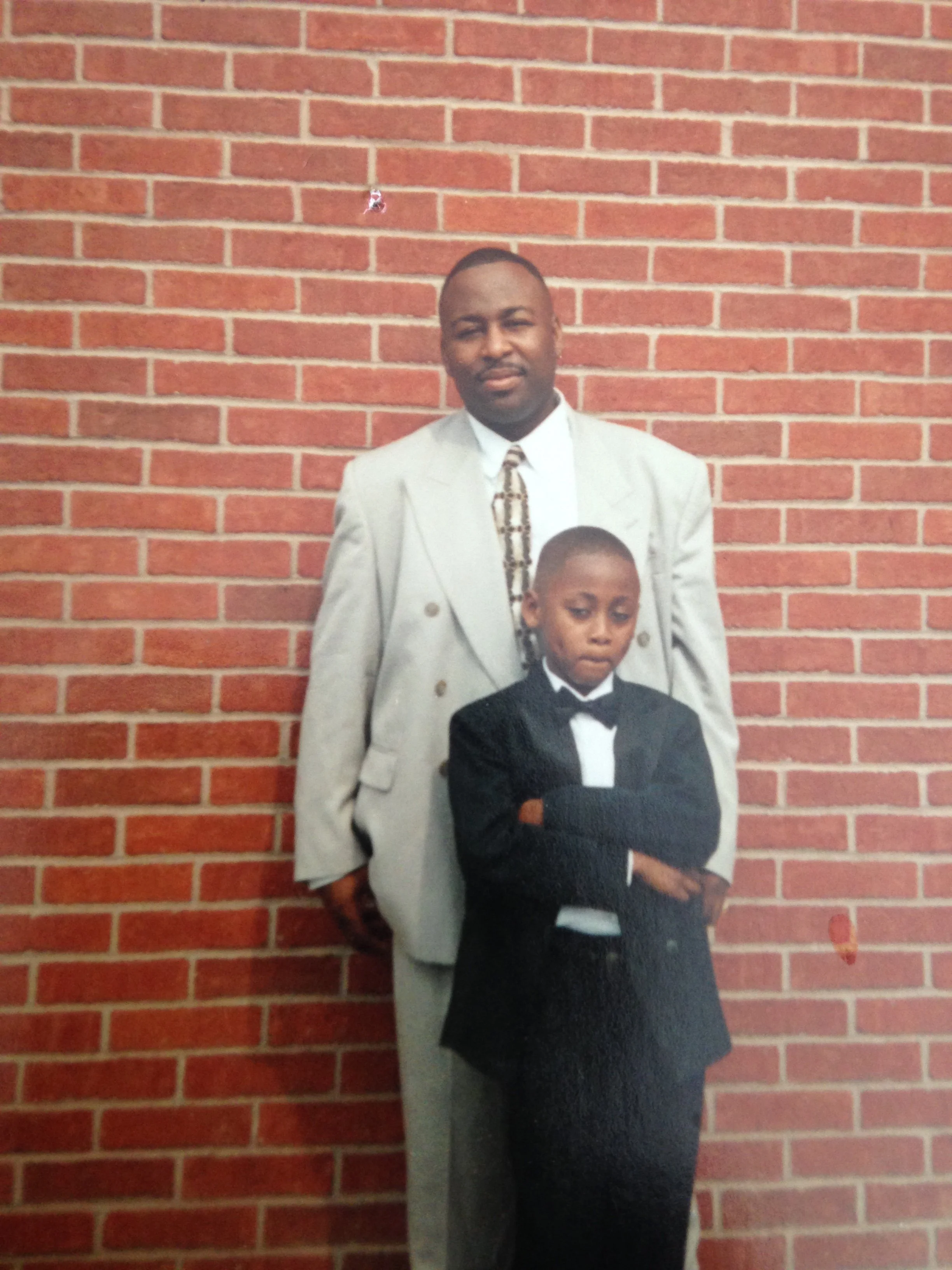 A man and a boy standing in front of a red brick wall. The man is dressed in a light-colored suit with a patterned tie, and the boy is wearing a black tuxedo with a bowtie, arms crossed.