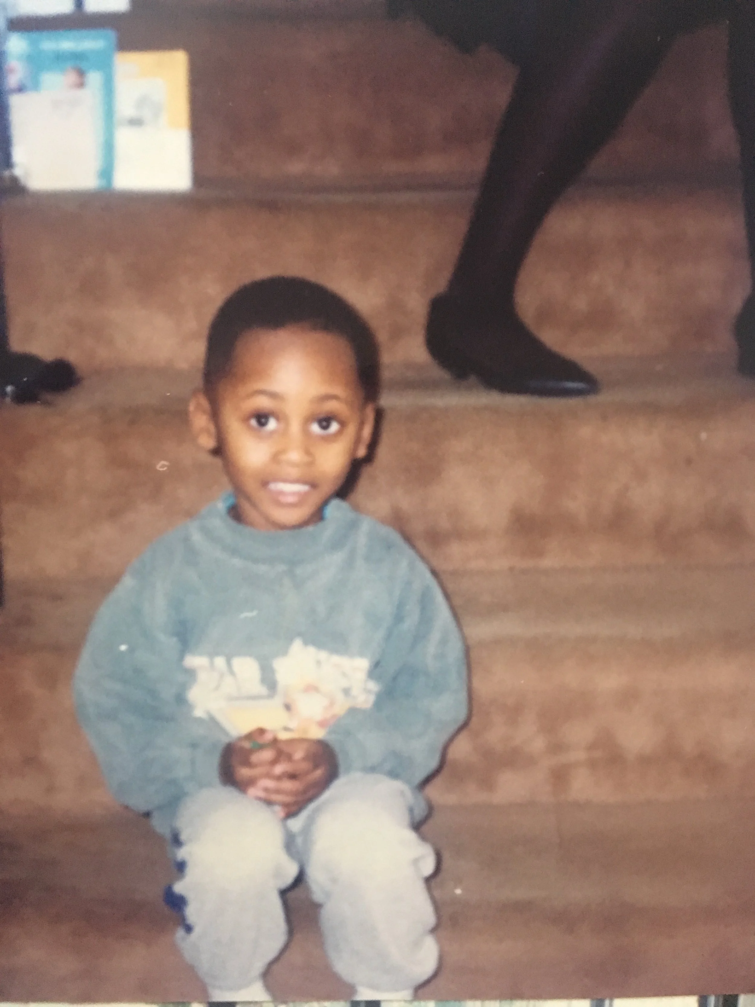 A young boy with short hair, wearing a gray sweatshirt and light-colored pants, sitting on wooden stairs, smiling at the camera, with a person in black shoes and stockings walking in the background.