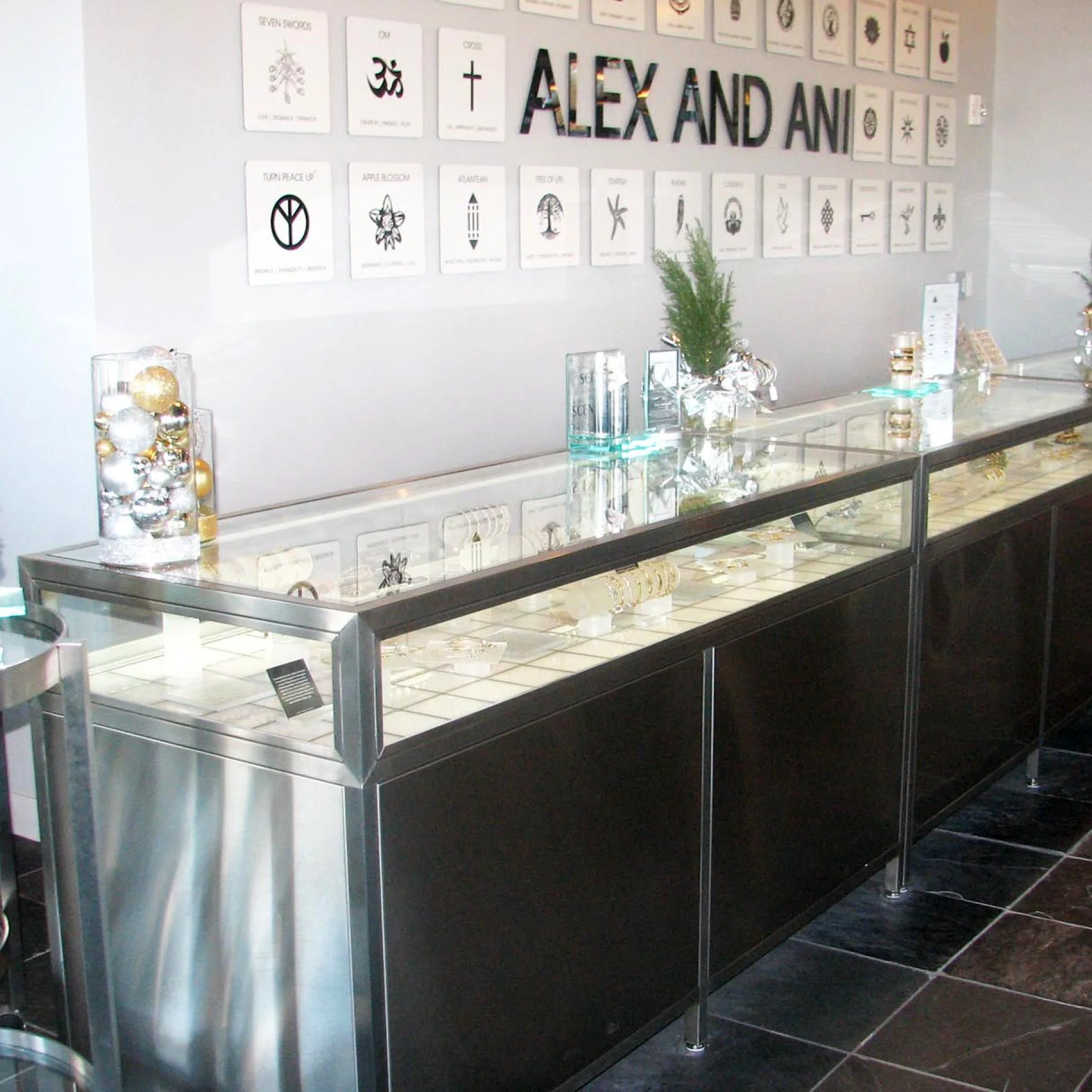 Display case with jewelry and holiday ornaments in a store, white wall with artwork and text, including "ALEX AND ANI", and a small green plant on the counter.