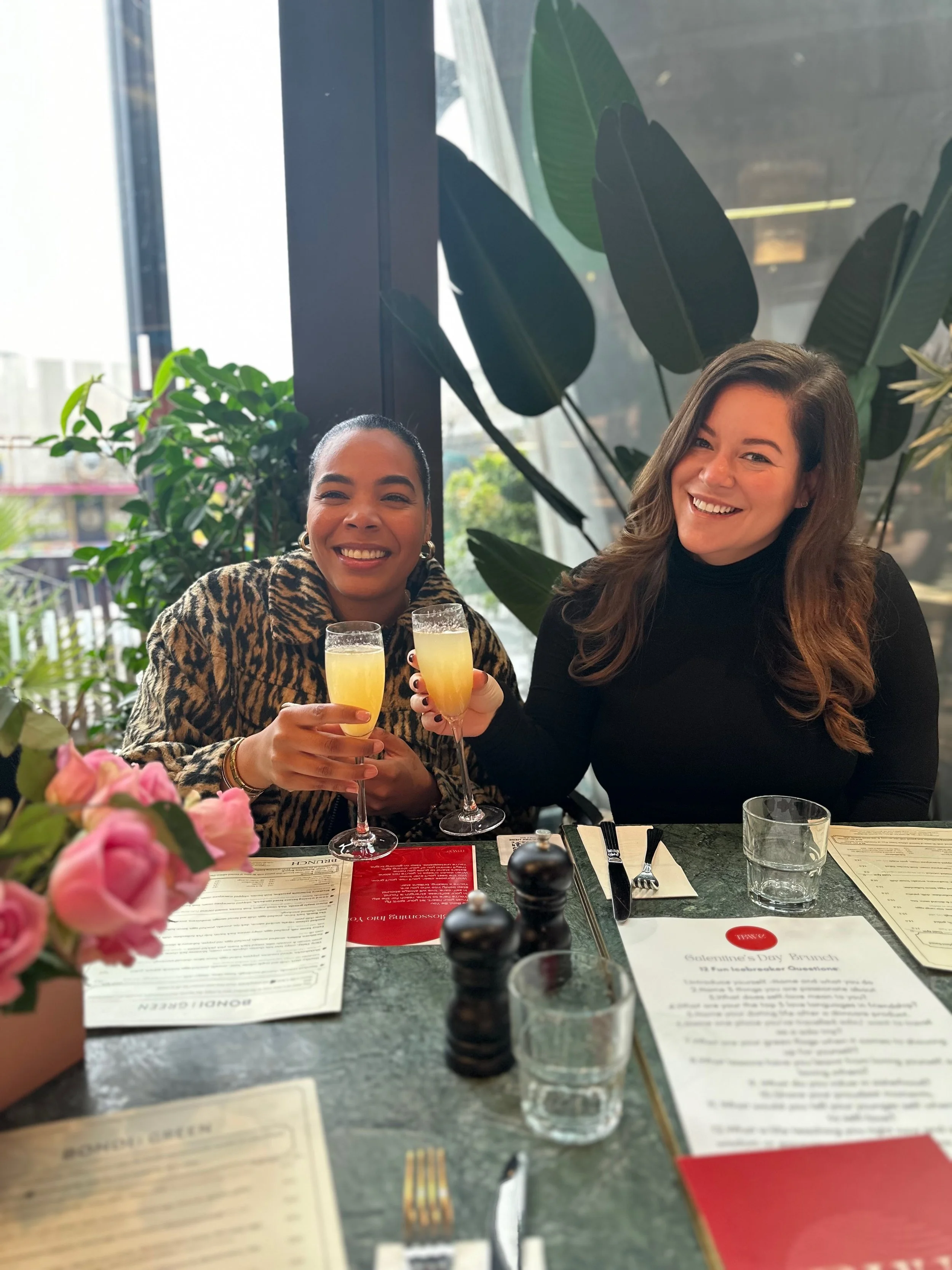 Two women sitting at a restaurant table, smiling and toasting with glasses of mimosa, surrounded by menus, flowers, and greenery, with a large glass window in the background.
