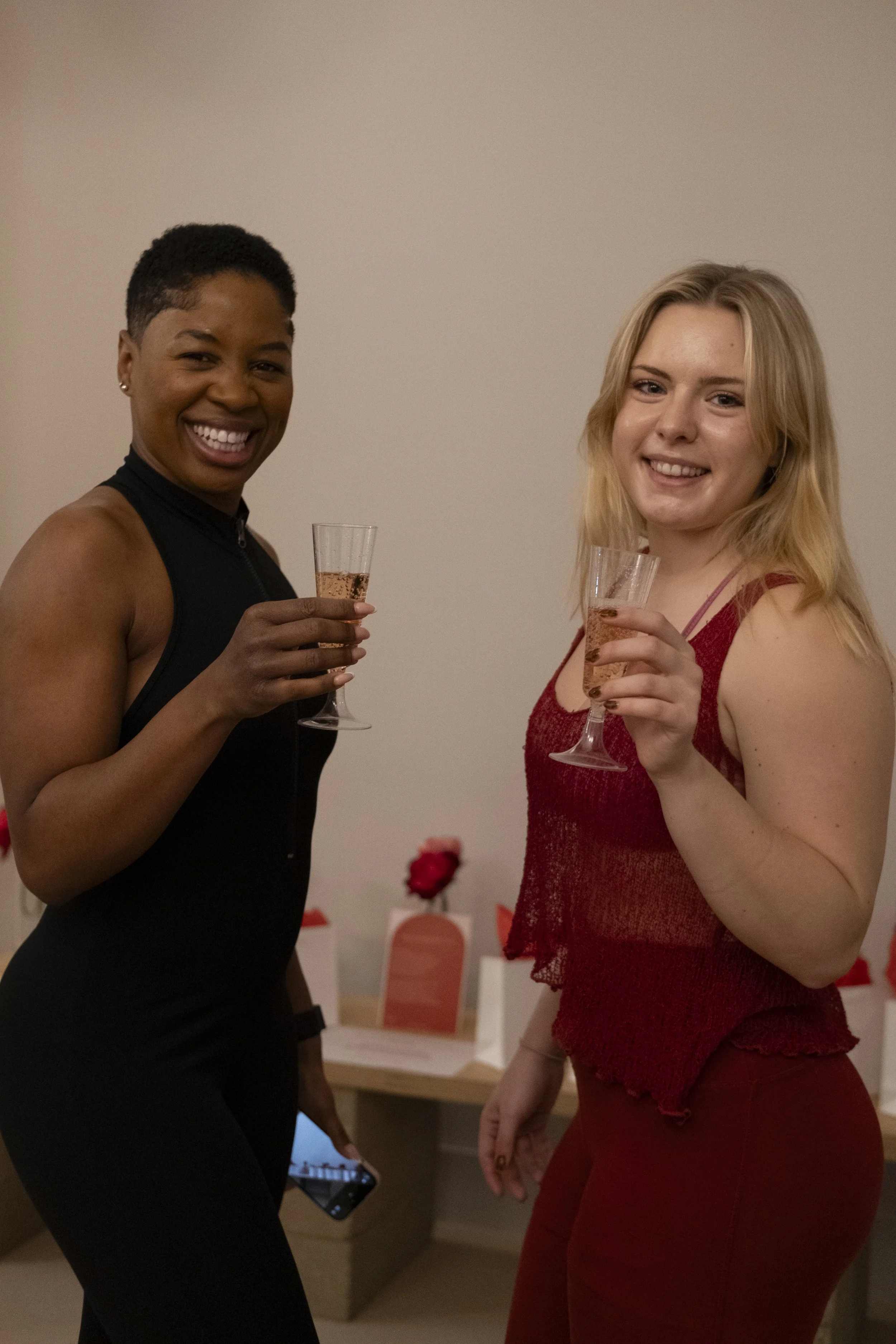 Two women celebrating with champagne glasses, one with dark skin and short hair in a black dress, the other with light skin and blonde hair in a red outfit, smiling at a social gathering.