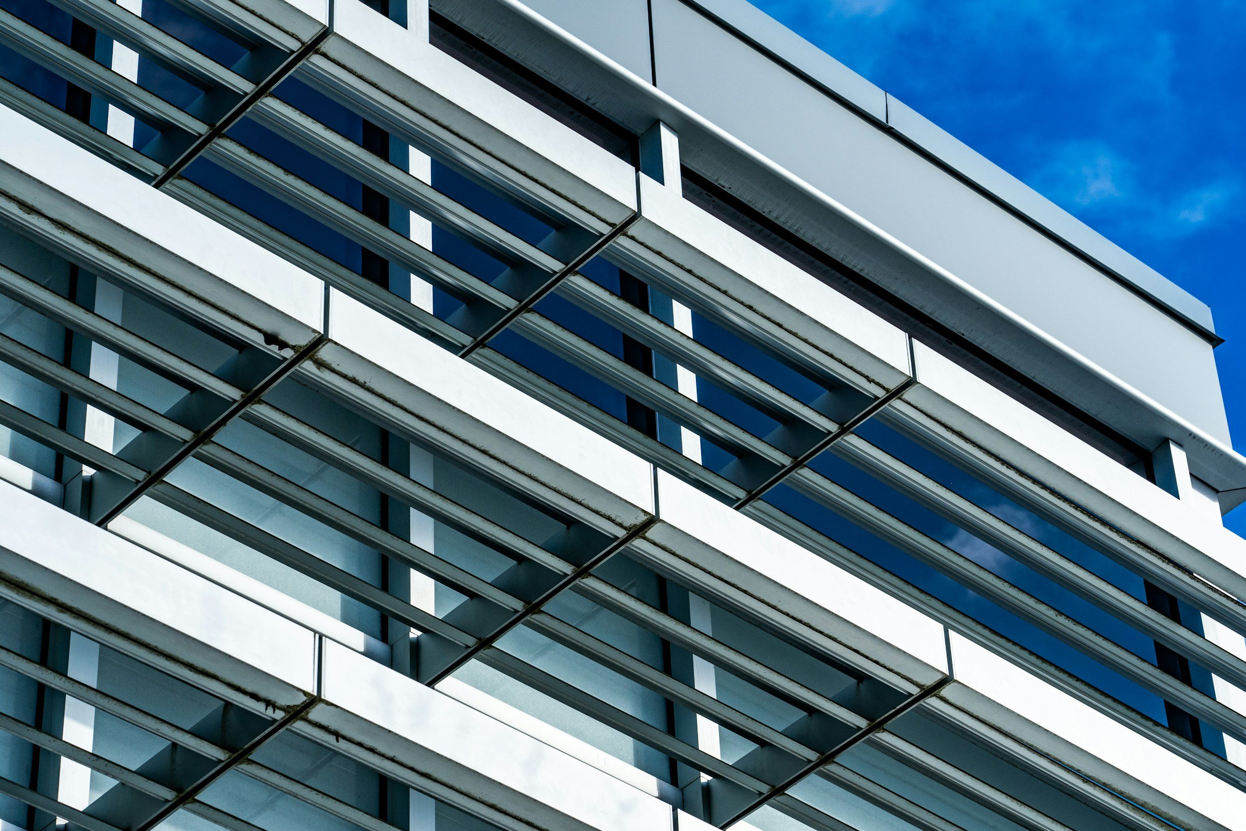 Close-up of a modern glass and metal building with horizontal white panels and blue-tinted windows, under a partly cloudy sky.
