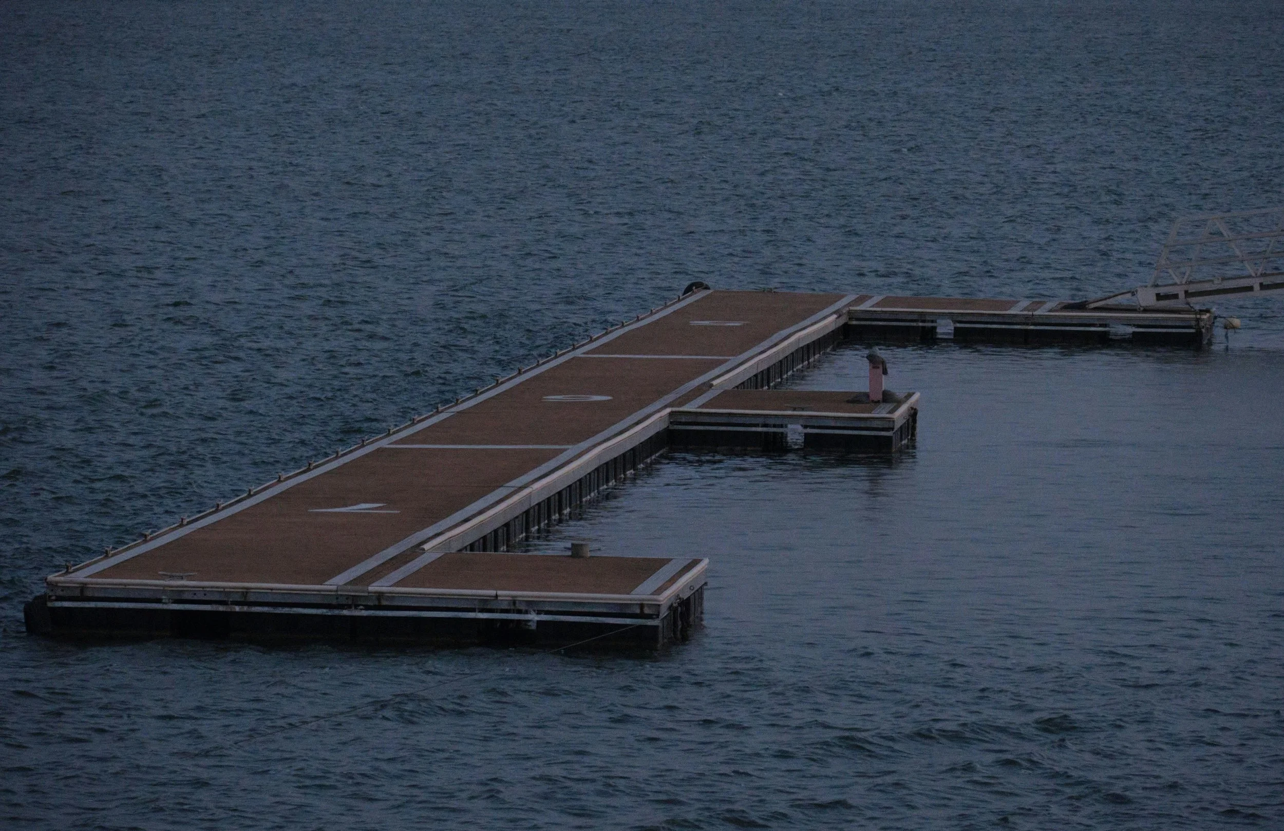 A wooden dock extending into a body of water with a floating section and a crane in the background.