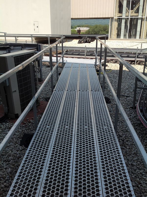 Metal grated walkway on rooftop with air conditioning units and industrial buildings in the background.