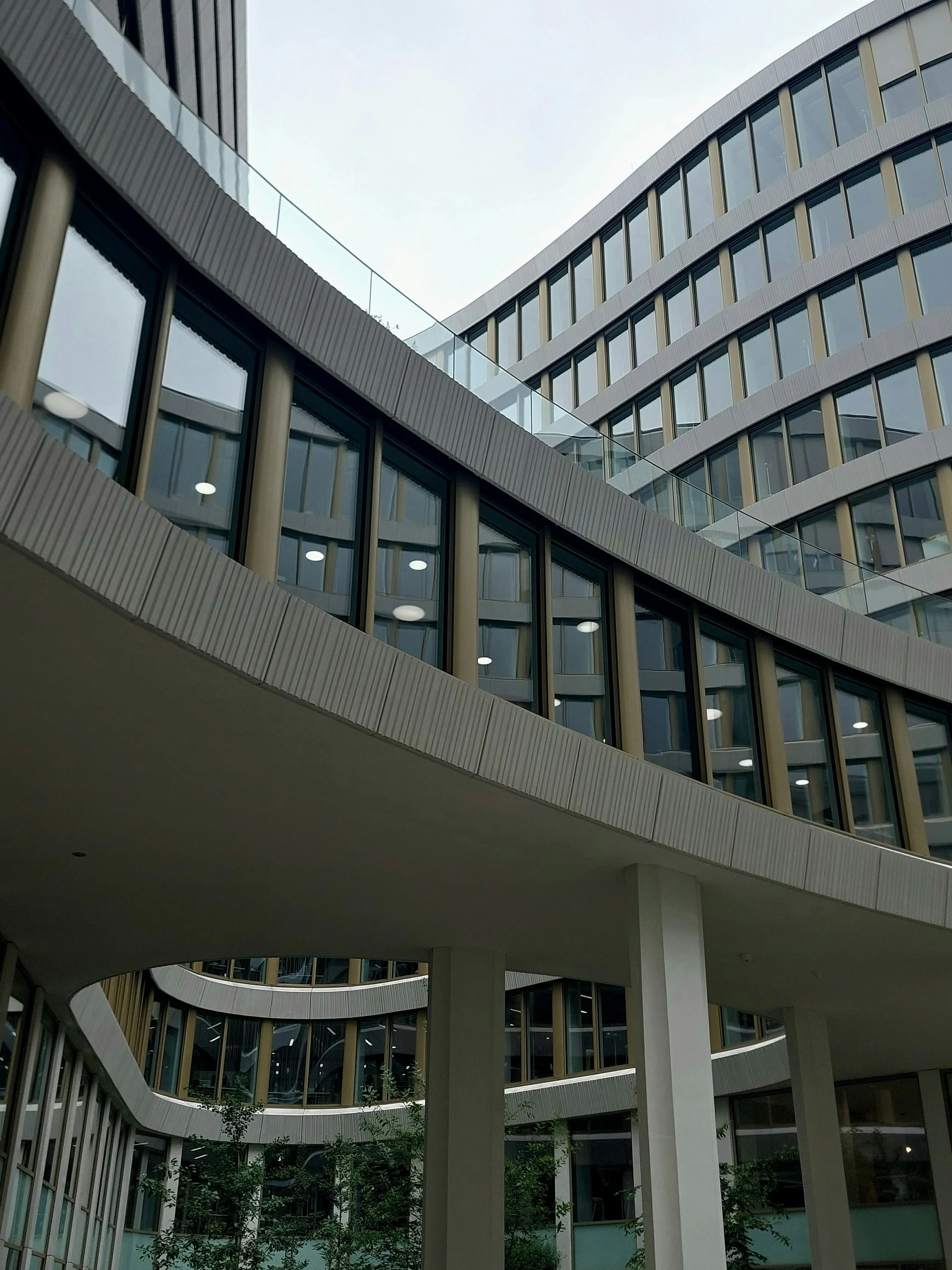 Modern building with large glass windows and curved architectural design, with greenery at the base and an overcast sky above.