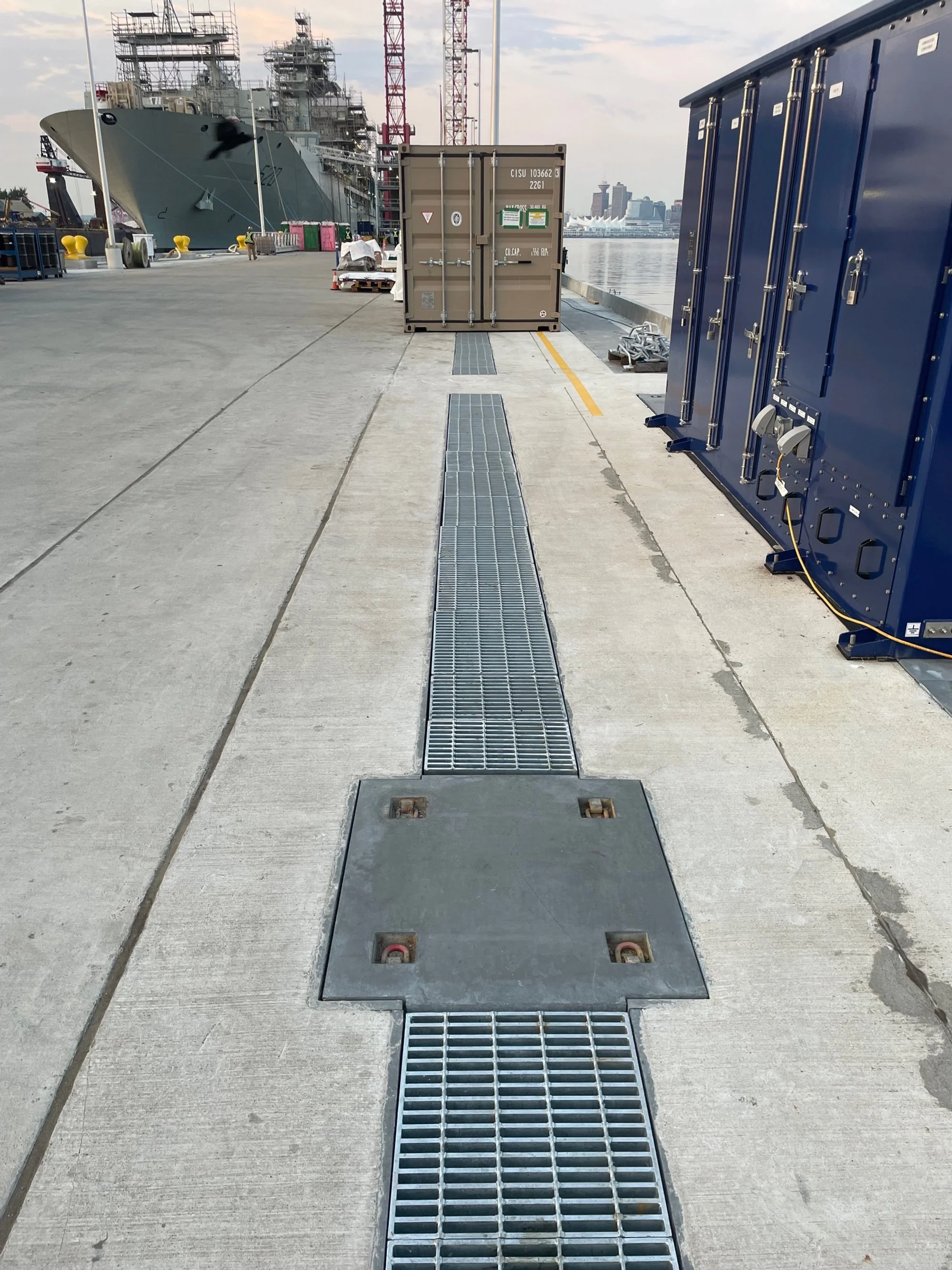 View of a ship docked at a harbor with various equipment and containers along the pier, including a large military vessel on the left, blue storage units on the right, and a city skyline in the background.