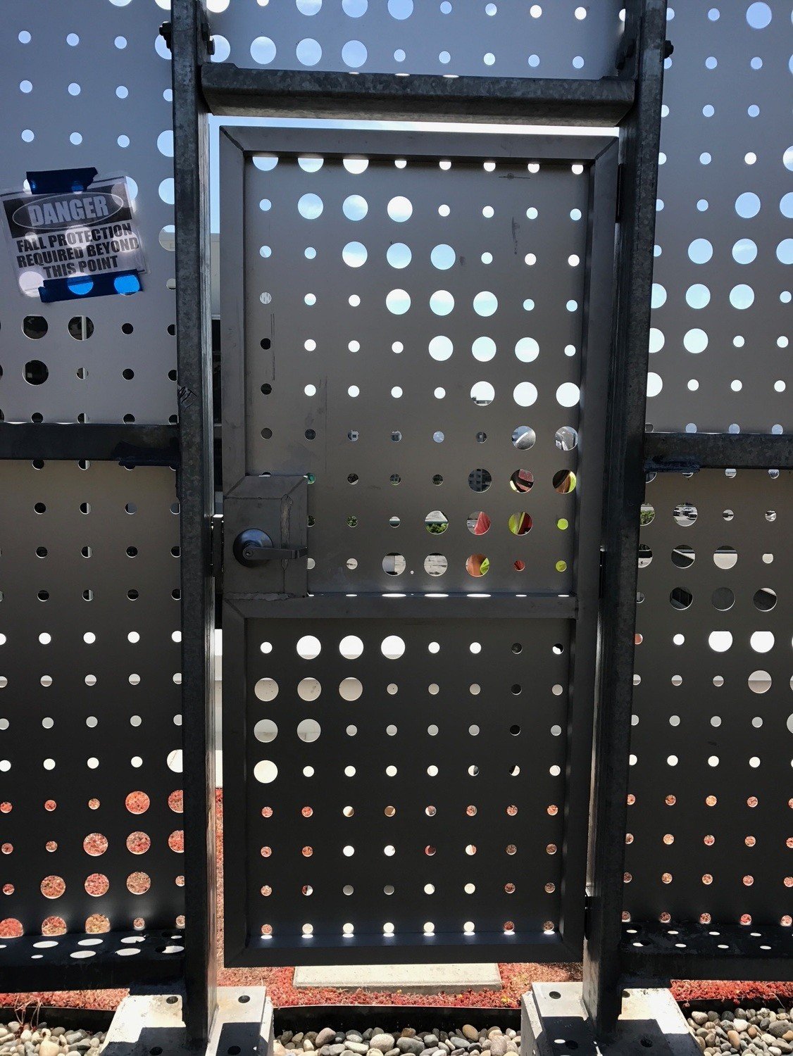 Metal door with a handle, set in a perforated metal wall with circular holes, and a warning sign on the left side that reads 'DANGER FALL PROTECTION REQUIRED BEYOND THIS POINT'.