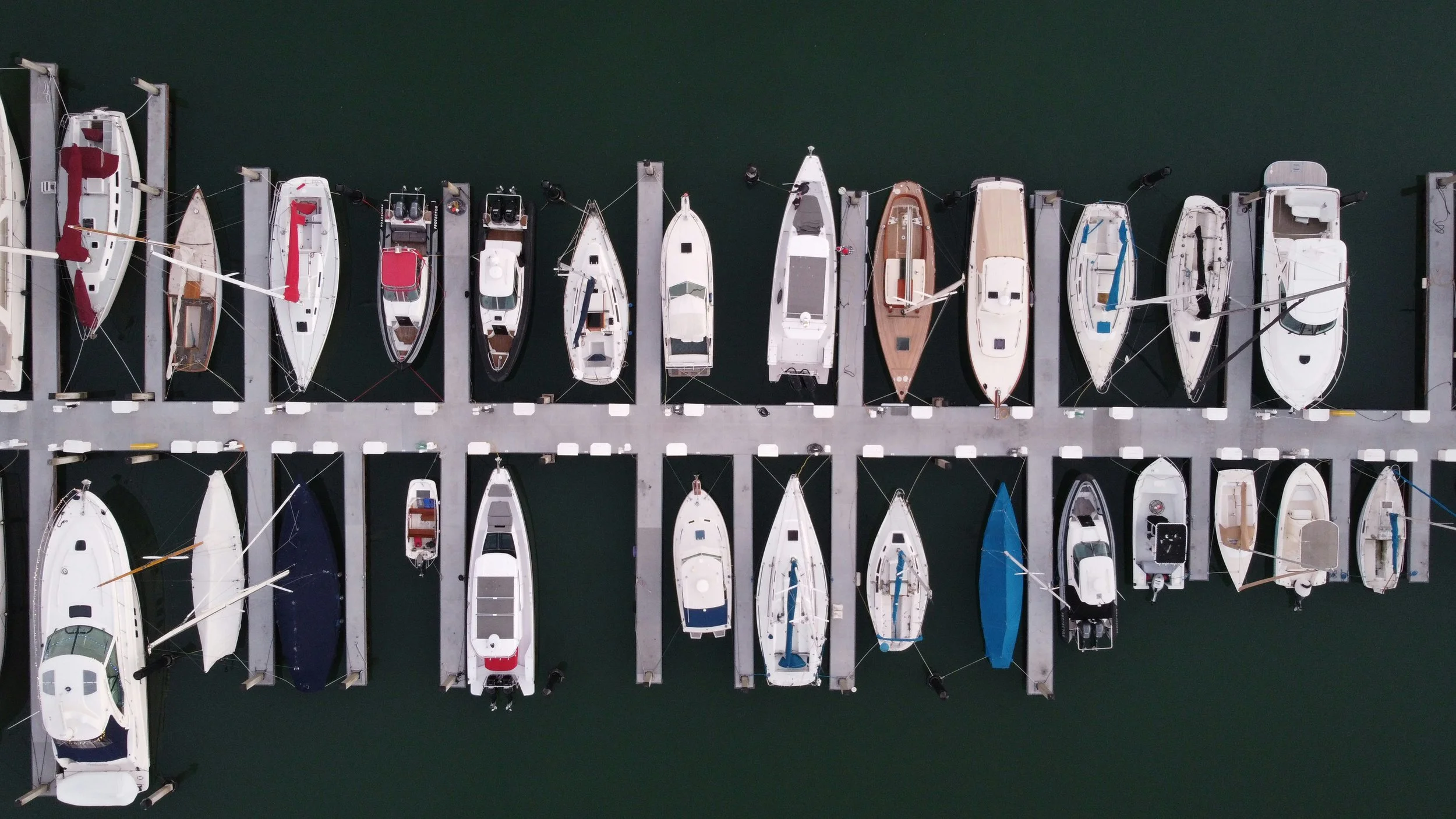 An aerial view of a marina with boats docked along two parallel piers extending into calm, dark water.