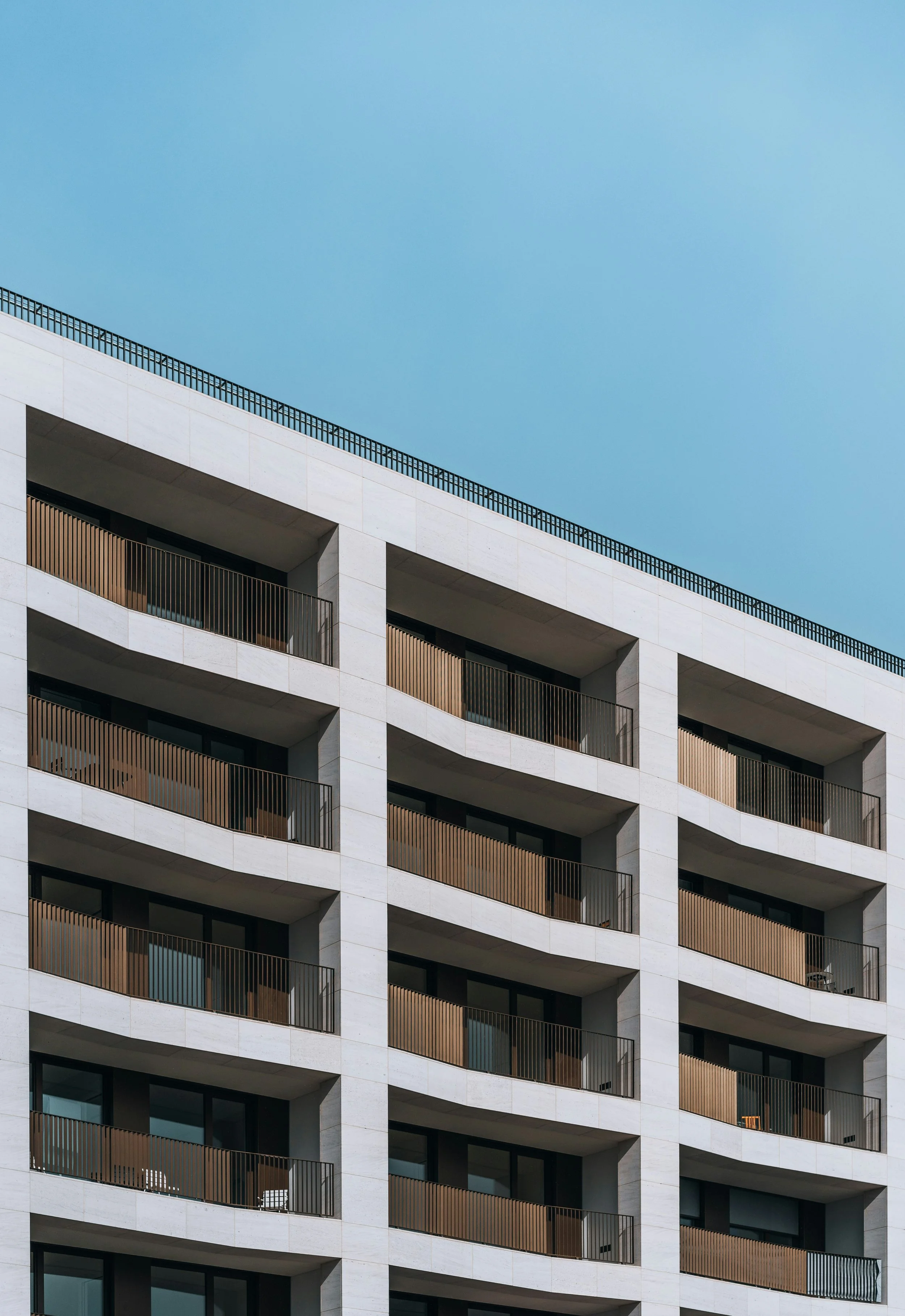 Modern multi-story apartment building with white exterior and wooden balcony railings under a blue sky.