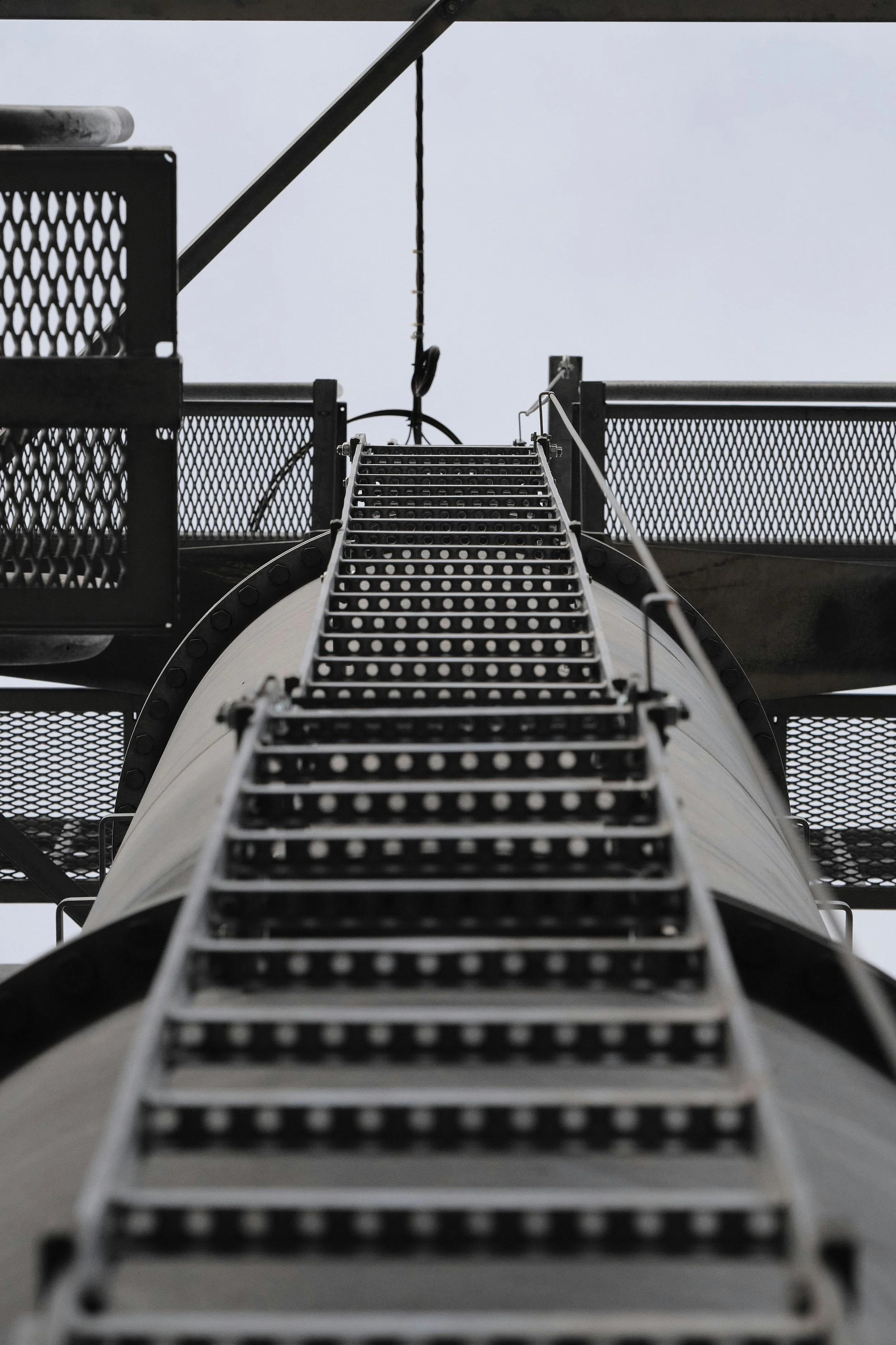 A metal ladder attached to a rocket or missile with surrounding metal platforms and guardrails, viewed from below against a cloudy sky.