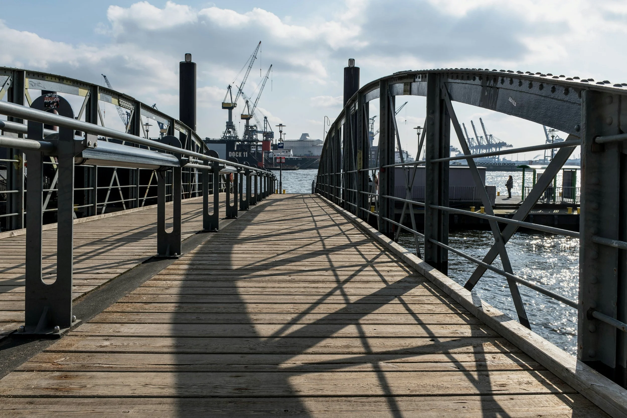 Wooden dock with metal railings leading to the water, with ships and cranes in the harbor and a woman walking in the distance on a cloudy day.