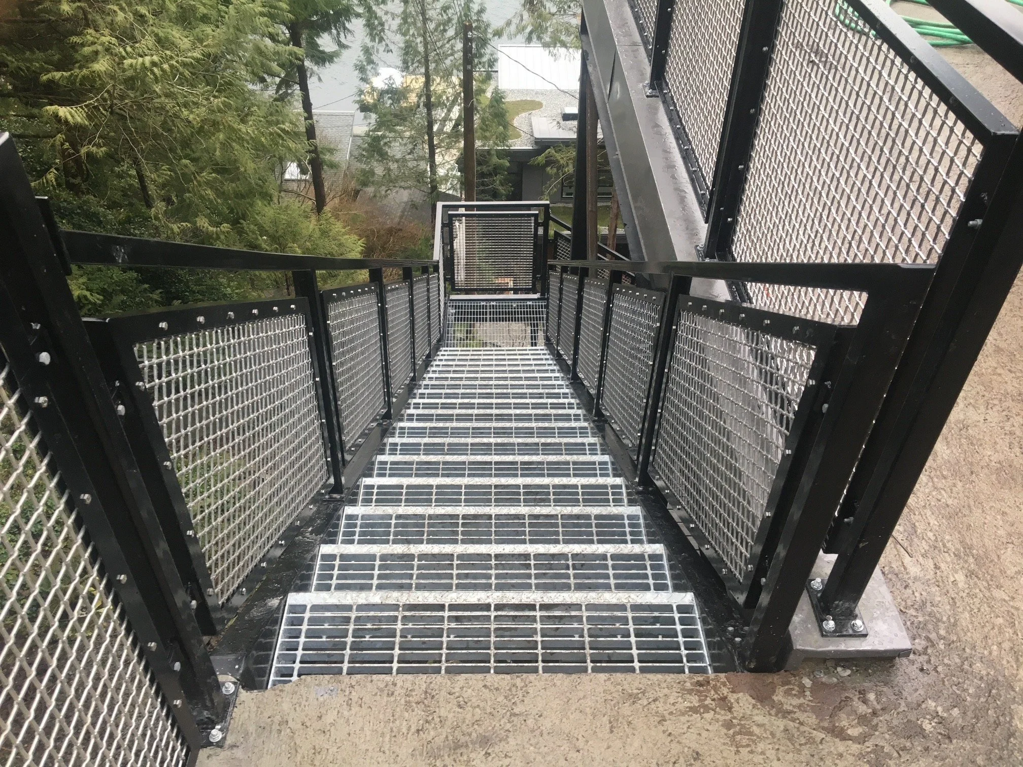 A metal staircase with black frame and safety netted sides, leading downward with a forested area in the background.
