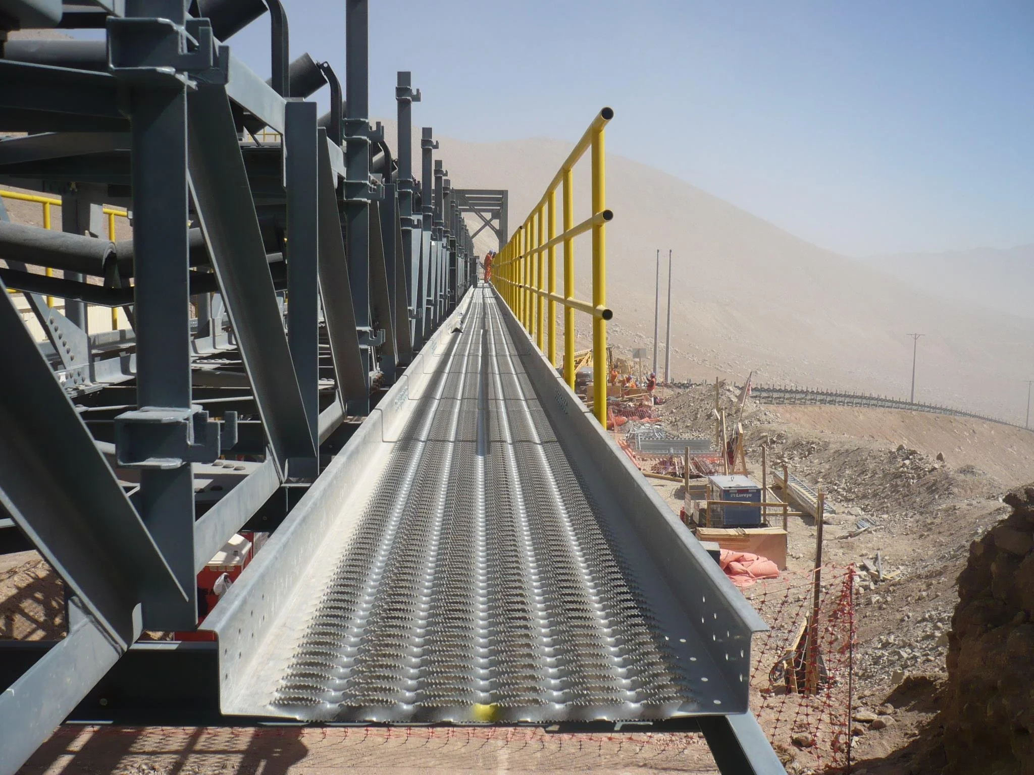 View of a construction site in a desert, featuring a metal walkway with yellow safety railings, construction equipment, and a barren landscape with mountains in the distance.