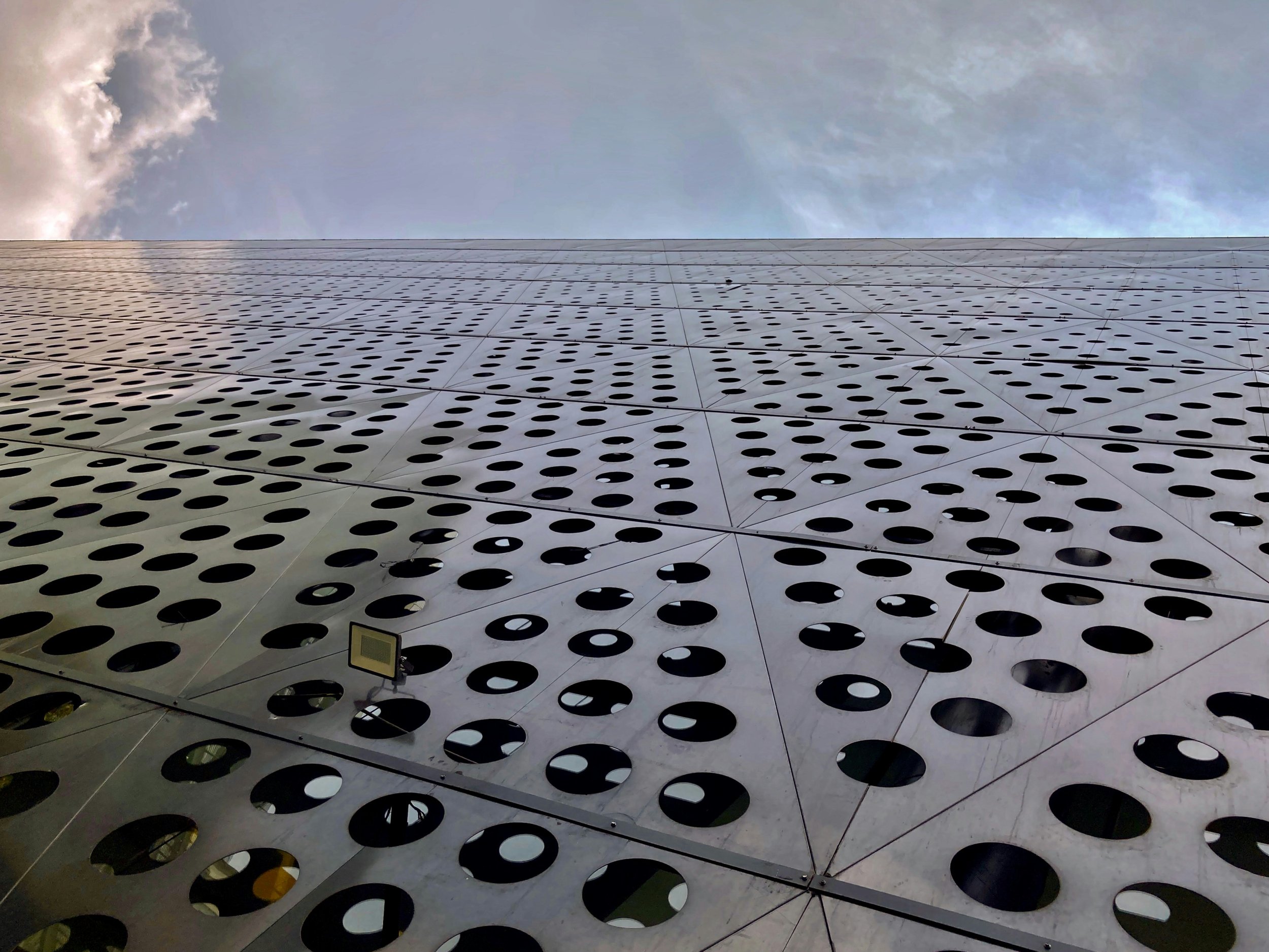 Metal staircase with black frame and silver grated steps, surrounded by metal mesh safety railings, leading downward with trees and buildings in the background.