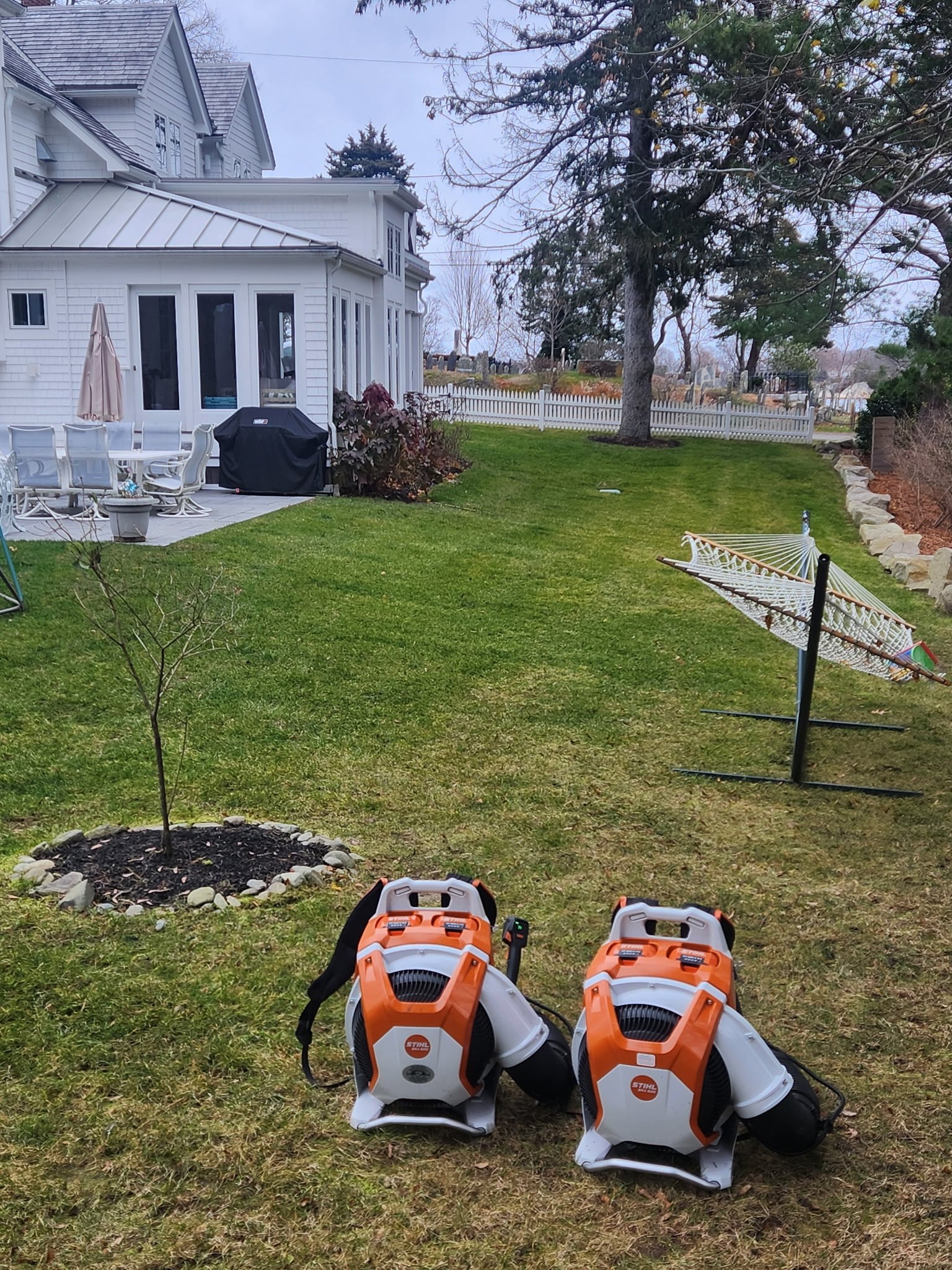 Backyard with a white house, outdoor patio furniture, a barbecue grill, a small tree with rock border, grass, a hammock, and two orange and white leaf blowers.