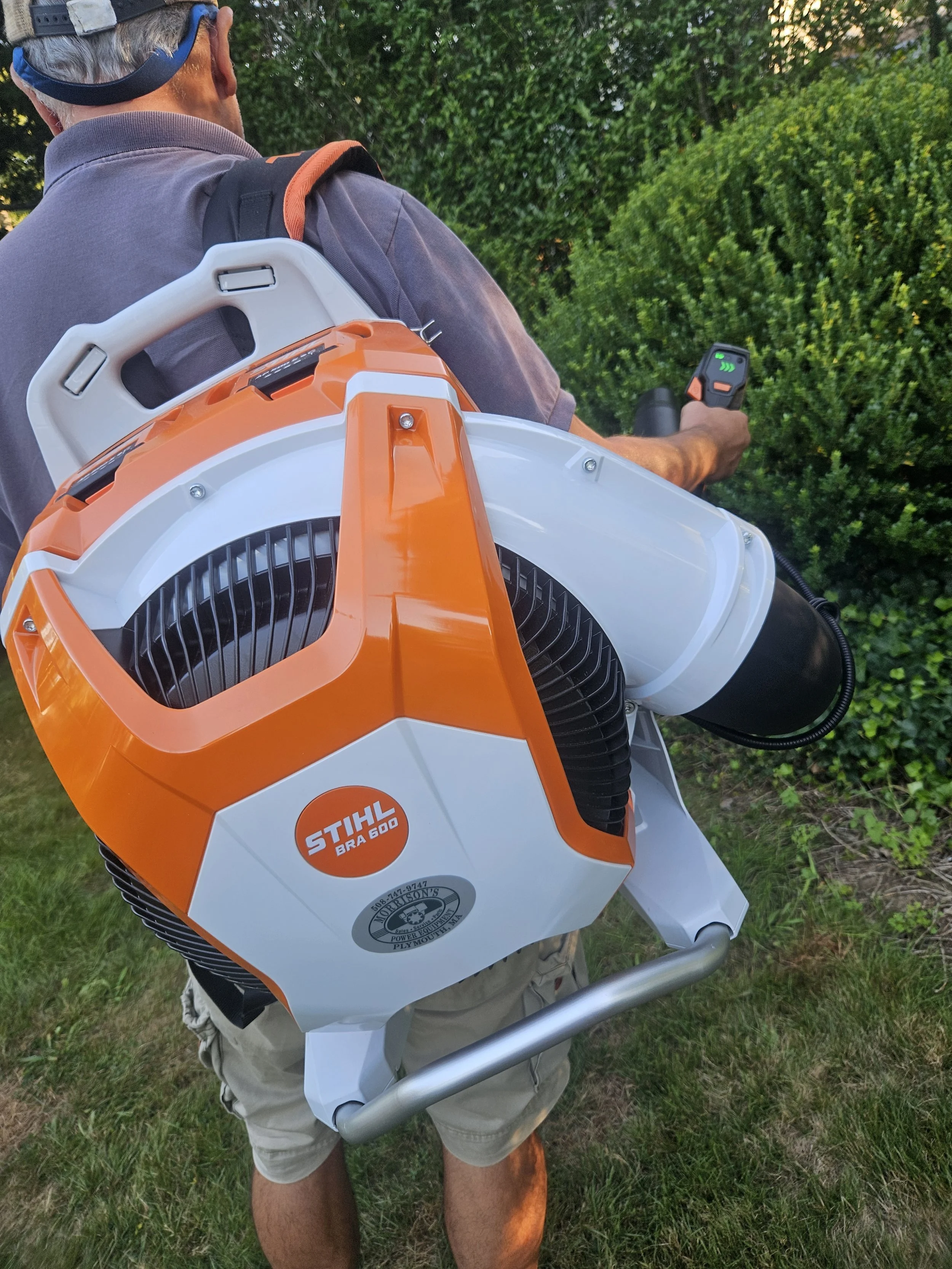 A person wearing a gray shirt, beige shorts, and a cap is using a battery-powered leaf blower with an orange and white body marked 'STIHL BR 600' to blow leaves or debris in a garden with green bushes and grass.