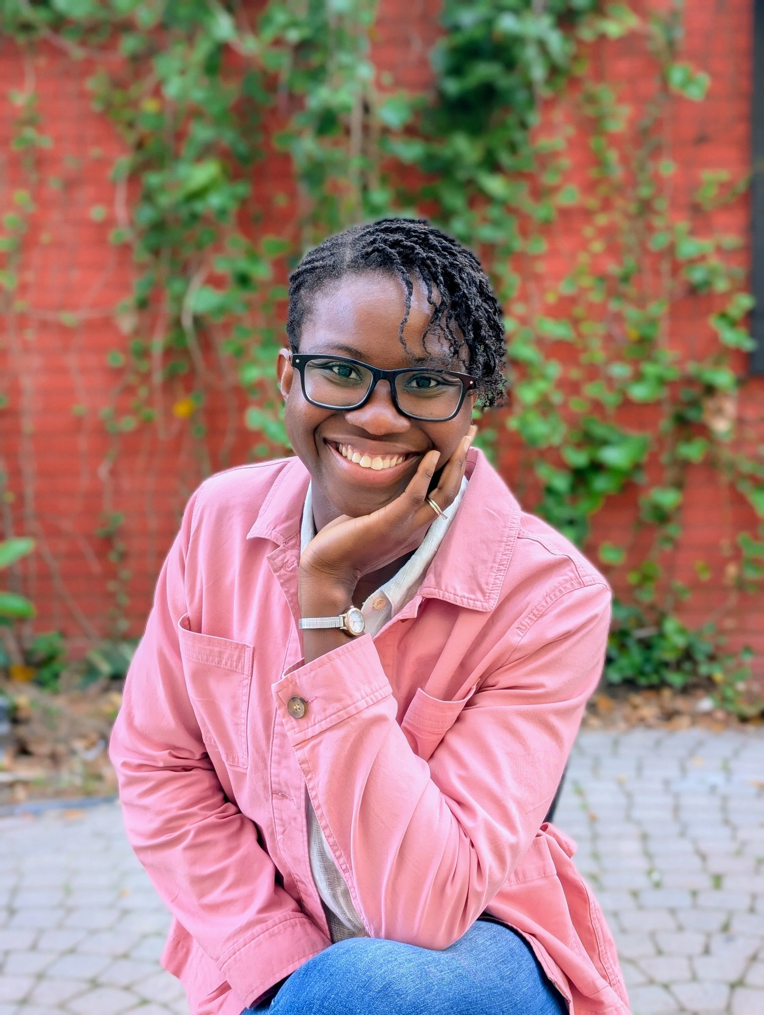 A smiling woman sitting outdoors against a red brick wall covered in wall crawlers  | Professional Organizer and Life Coach | Household Stewardship LLC