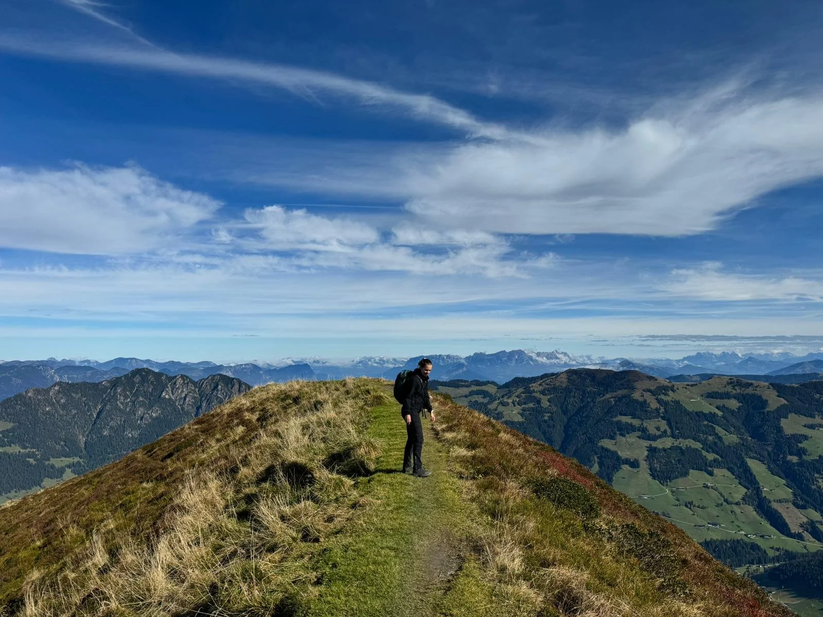Person auf einem Pfad auf einem Berggipfel mit Blick auf eine bewaldete Berglandschaft und Himmel mit Wolken.