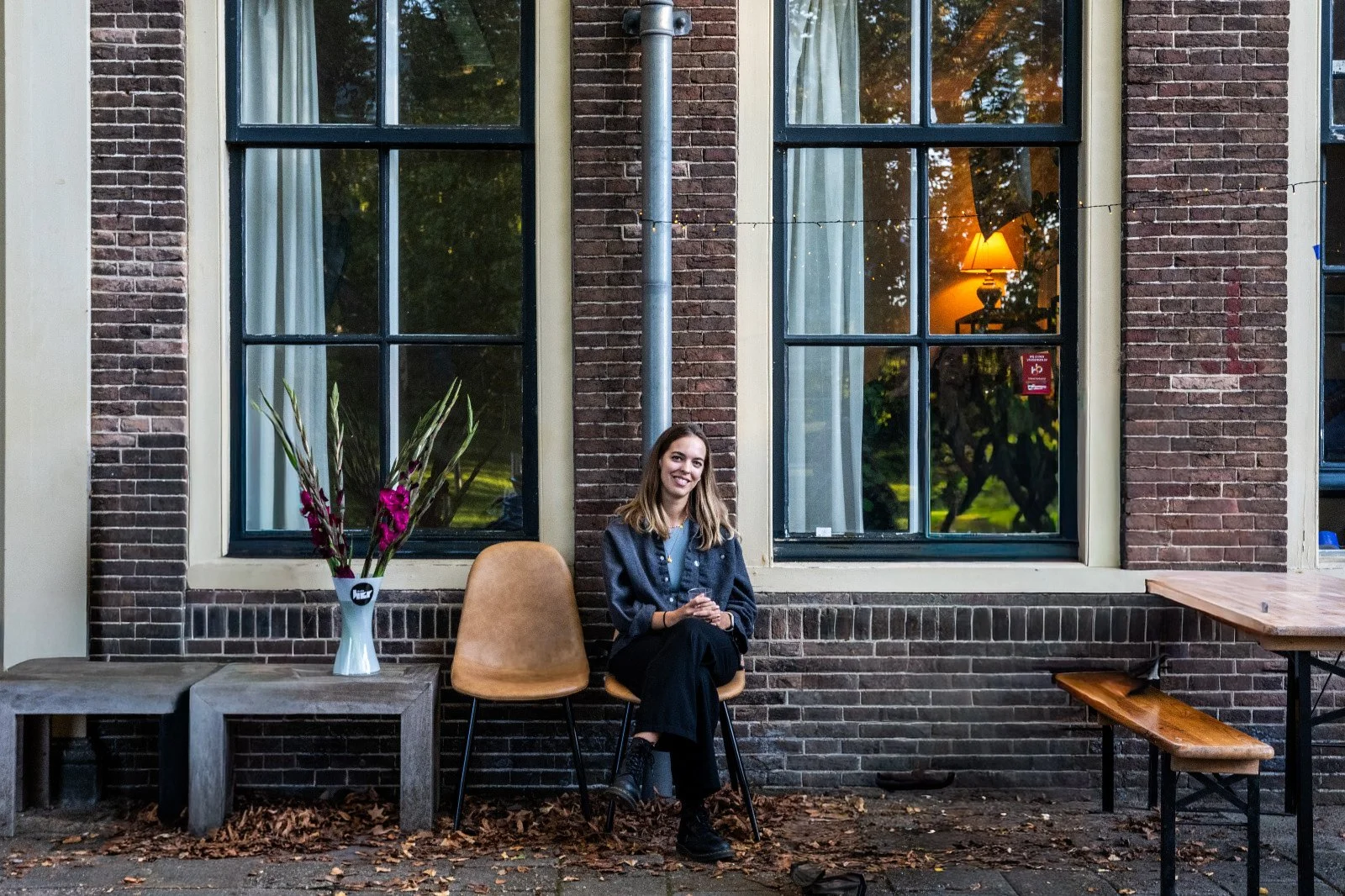 Eine junge Frau sitzt entspannt auf einem Stuhl vor einer Ziegelwand mit großen Fenstern, innen ist ein Tisch mit einer Lampe sichtbar, draußen liegen Herbstblätter auf dem Boden.