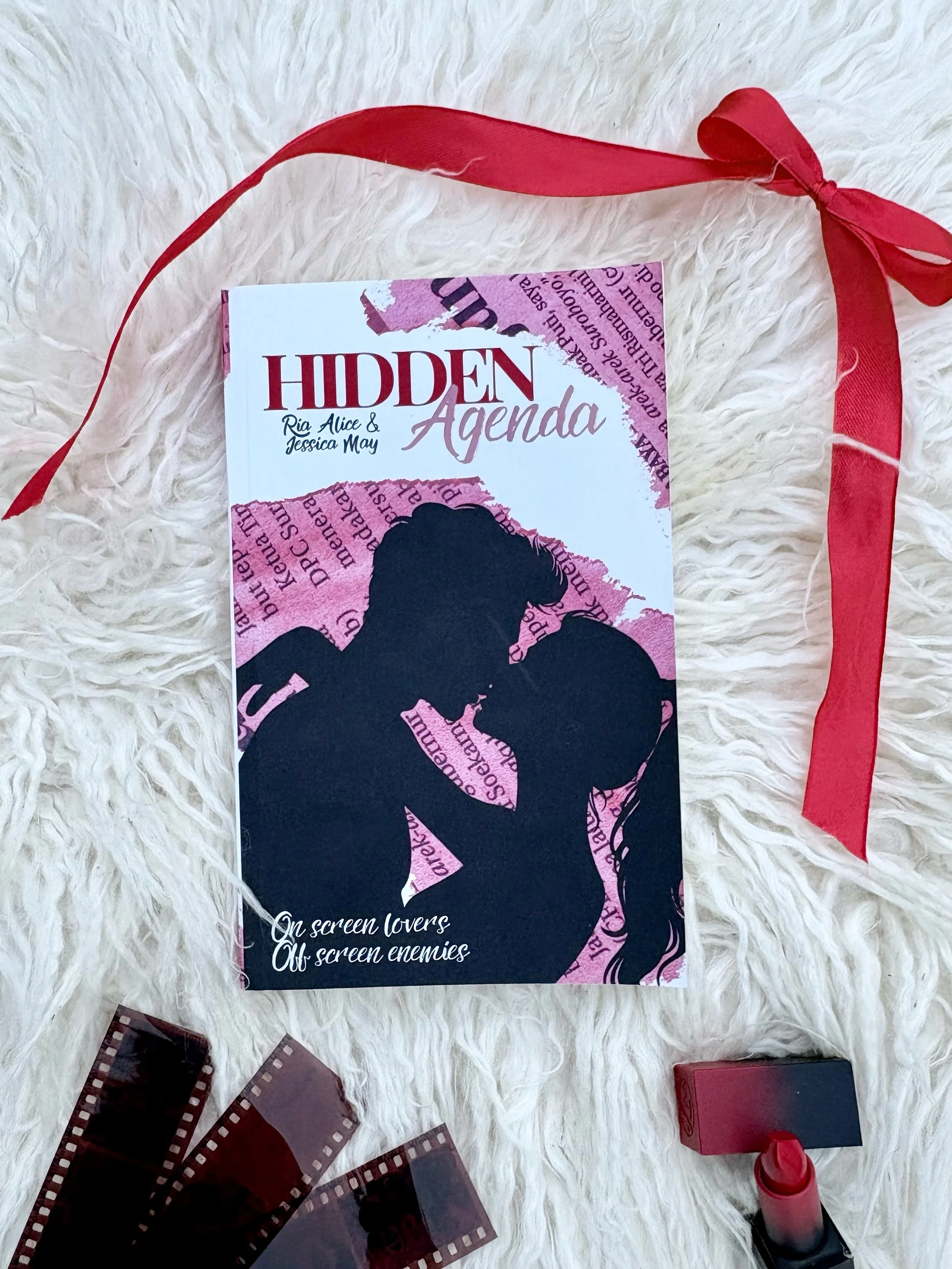 A book titled "Hidden Agenda" by Ria Alice and Jessica May is on a white, fluffy surface, with a red ribbon, some film strips, and a lipstick nearby.