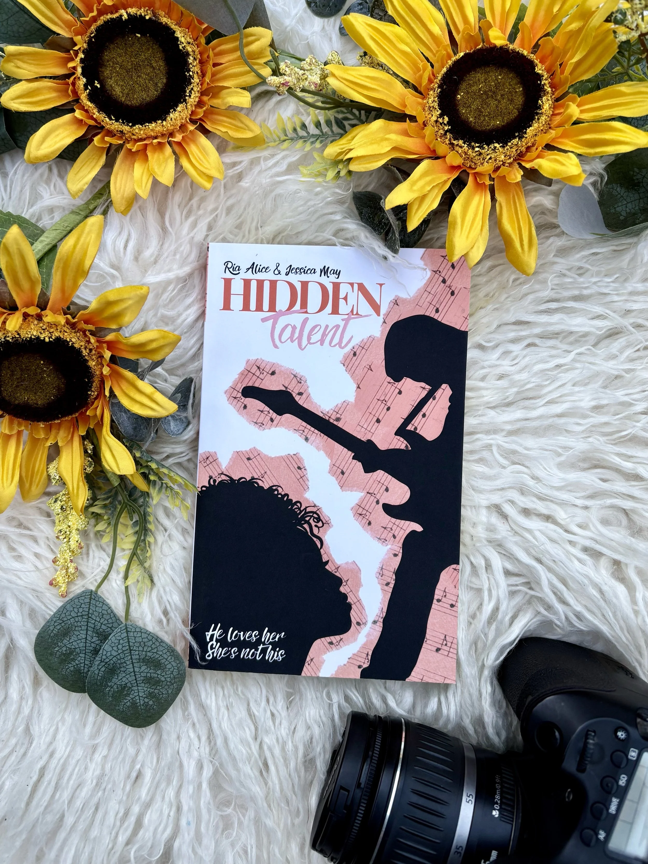A book titled "Hidden Talent" by Ria Alice and Jessica May, surrounded by yellow sunflowers, green leaves, and a camera on a fluffy white surface.