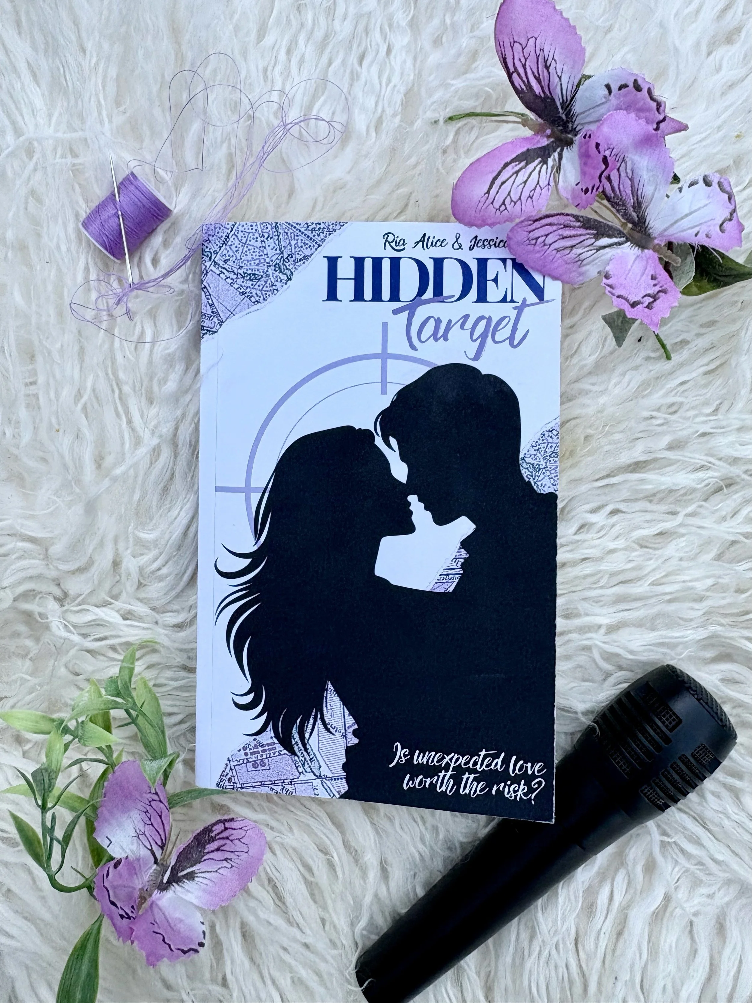 A romance novel titled 'Hidden Target' by Ria Alice and Jessica, with a silhouette of a man and woman about to kiss, placed on a soft white surface, surrounded by purple flowers, a spool of purple thread, and a black microphone.