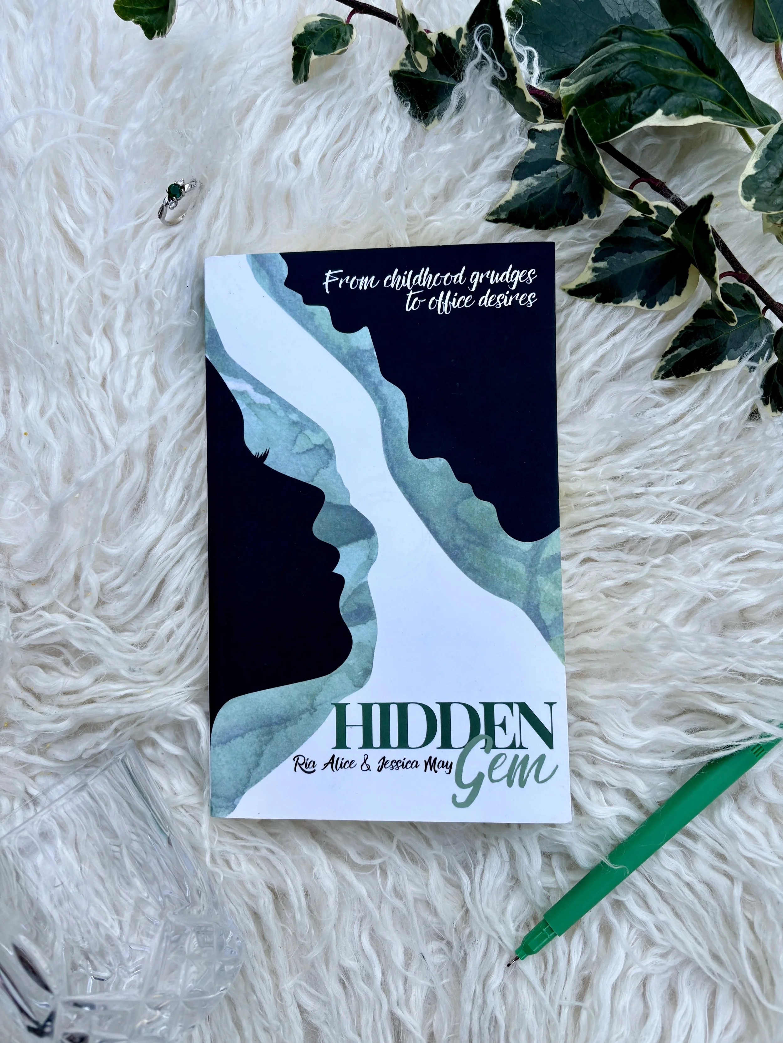 A book titled "Hidden Gem" by Ria Alice and Jessica May lies on a fluffy white surface, with a green pen, a glass, some jewelry, and green leaves surrounding it.