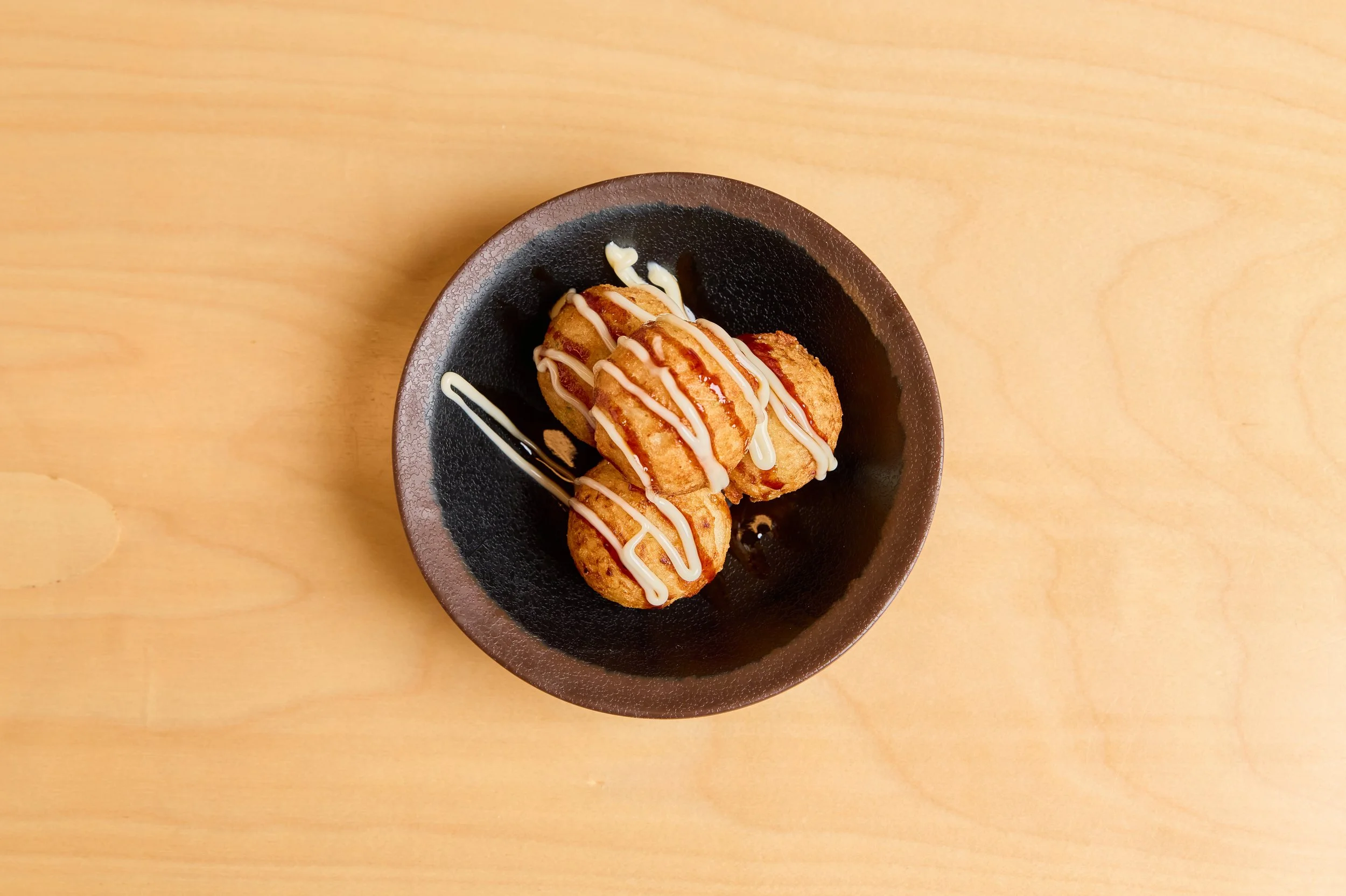 Três croquetes empanados decorados com molho branco e molho barbecue, servido em uma tigela preta sobre uma mesa de madeira clara.