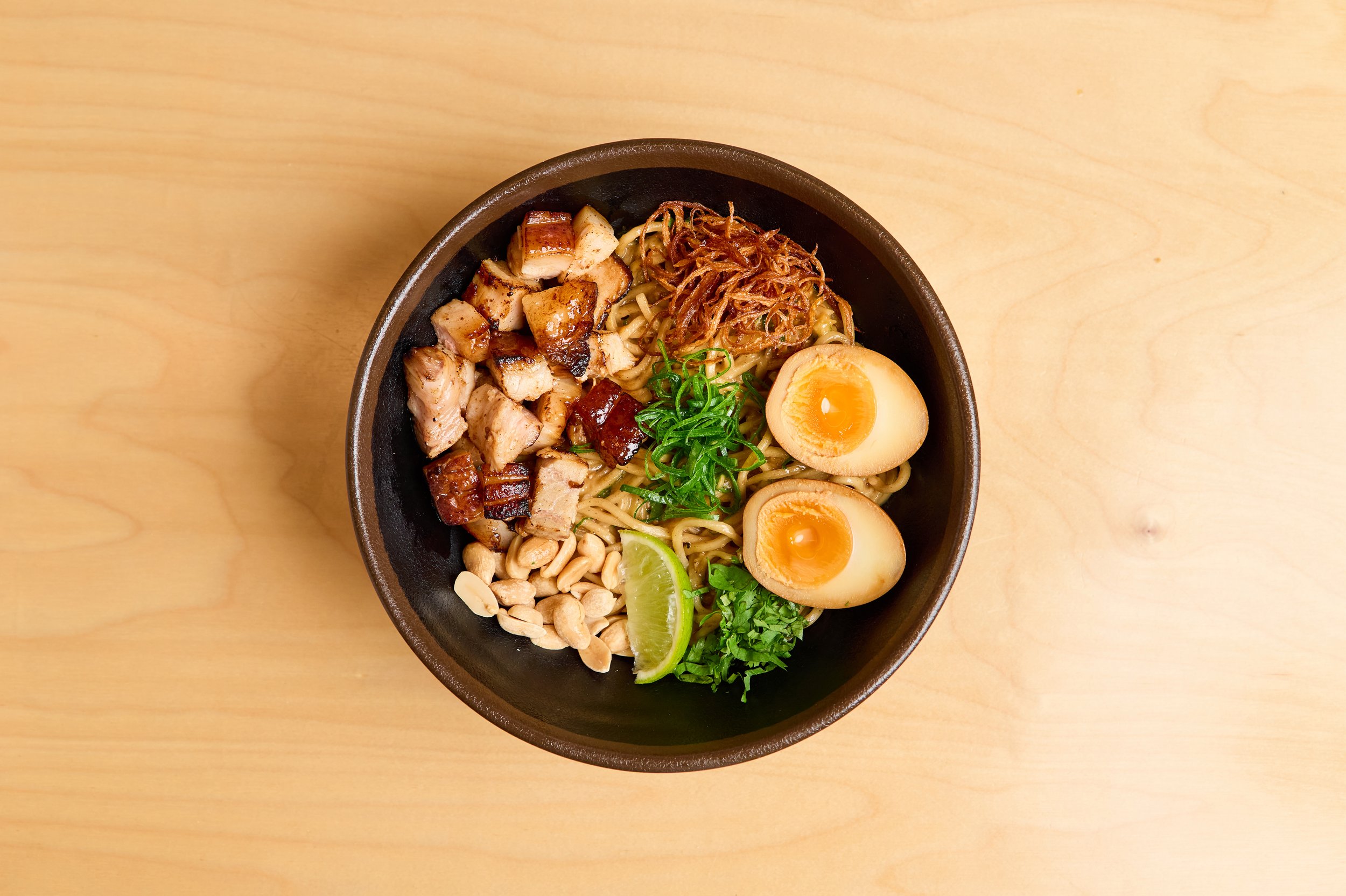 Tigela preta com ramen, fatias de ovo cozido, carne de porco, frango, caldo, cebolas fritas, limão, alho, cebolinha e coentro, sobre uma mesa de madeira clara.