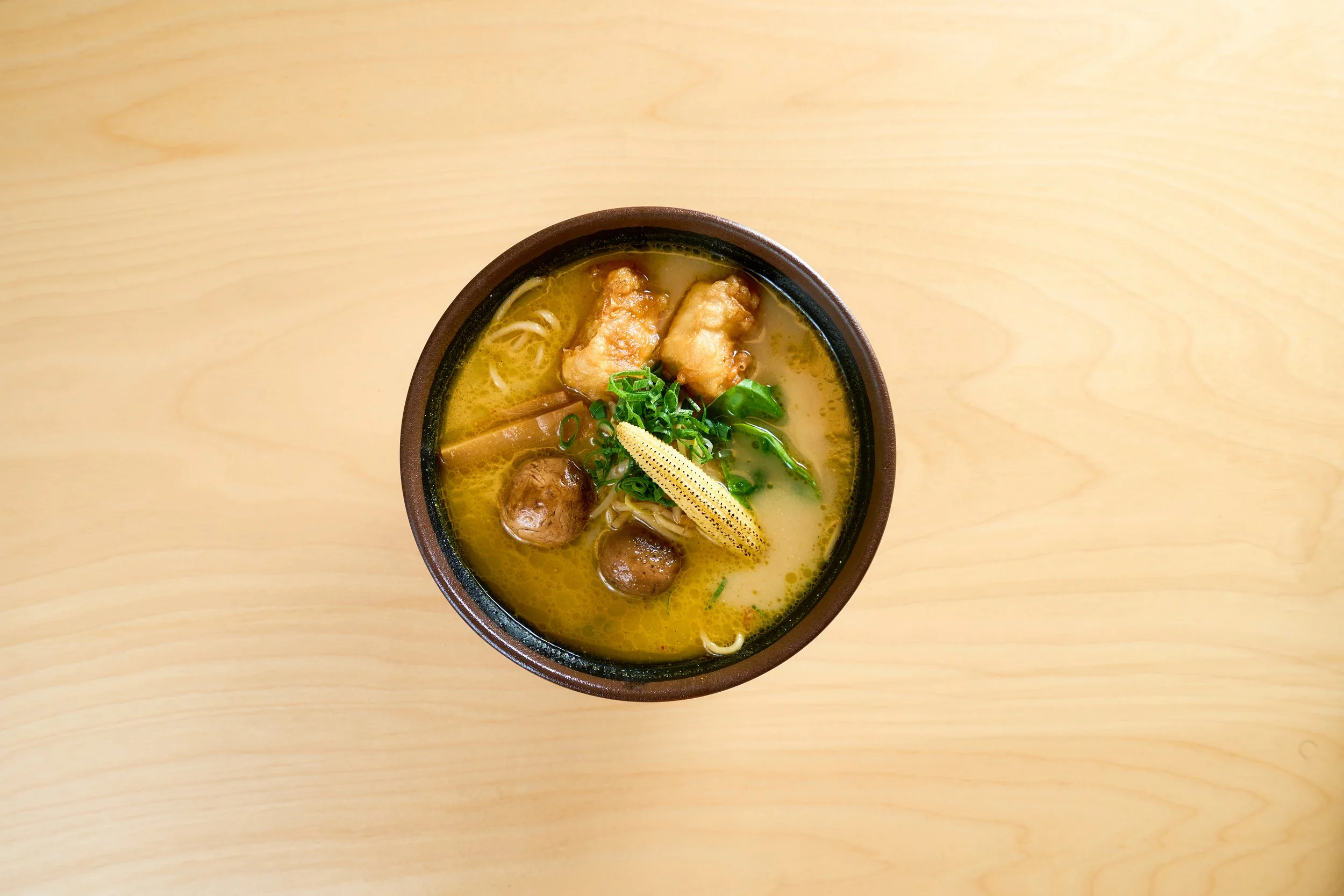 Uma tigela de sopa de ramen com caldo amarelo, pedaços de carne, bolinhos de carne, macarrão, cebolinhas verdes picadas, um pedaço de limão e um pedaço de baby corn, tudo sobre uma superfície de madeira clara.