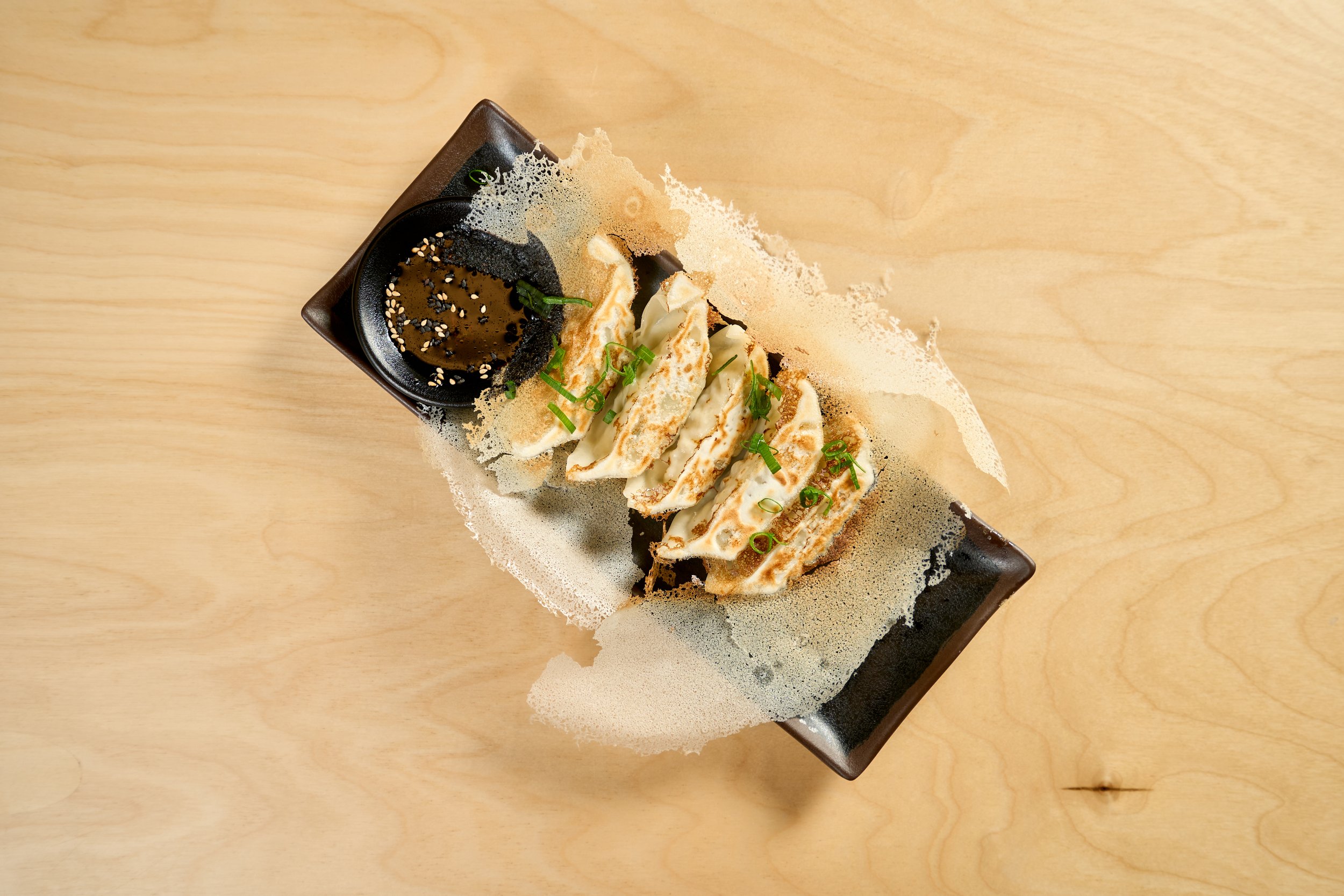 Porção de gyoza empanados com cebolinha e molho de soja com sementes de gergelim, servido em prato preto sobre mesa de madeira.