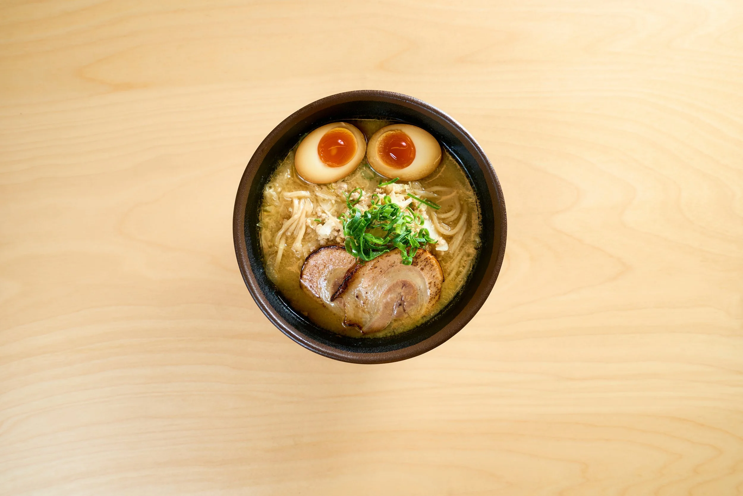 Tigela de ramen com ovos pochê, fatias de carne, macarrão, cebolinhas e caldo, sobre mesa de madeira